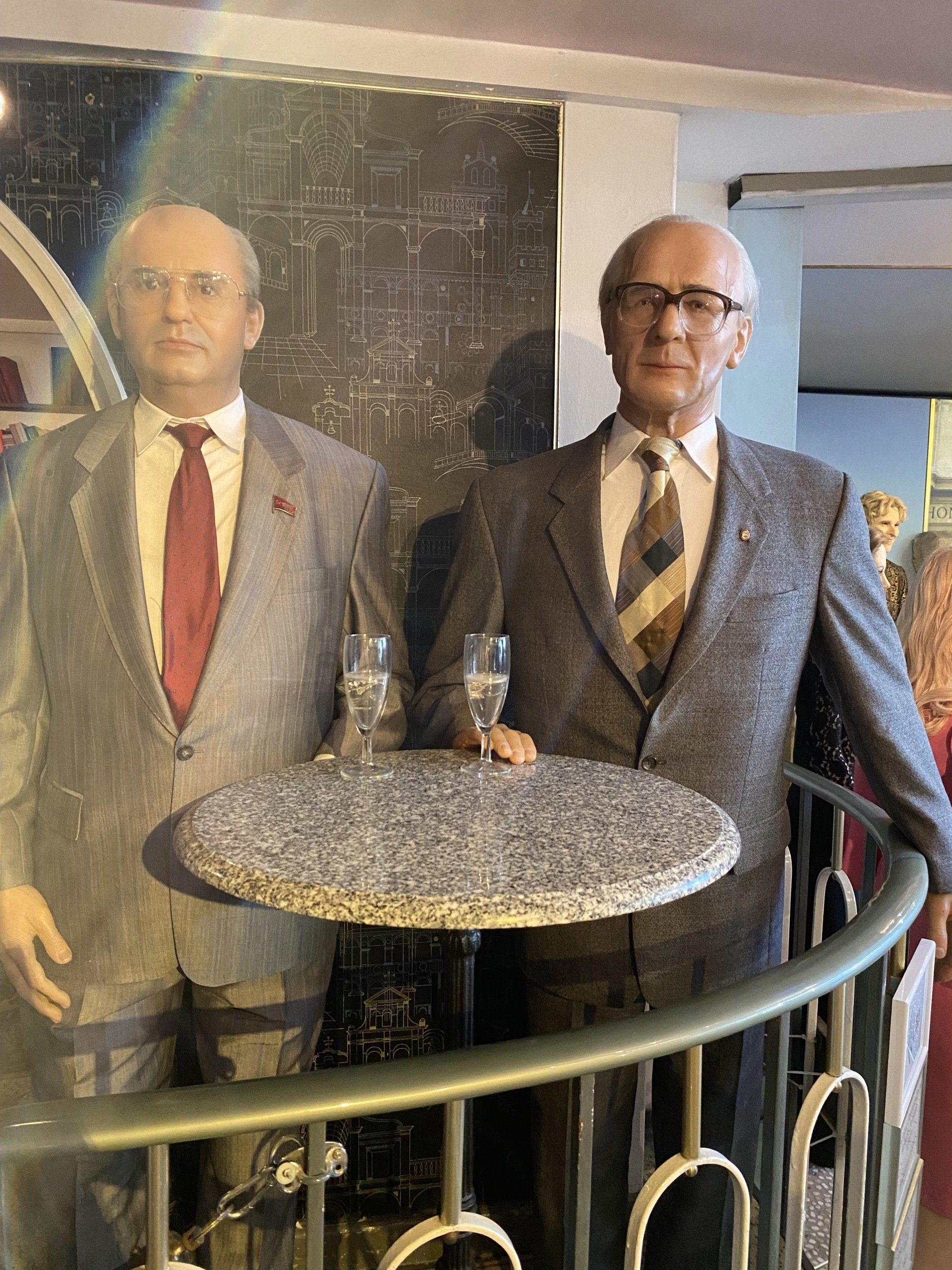 Wachsfiguren von zwei Männern in Anzügen an einem kleinen Tisch auf einem Balkon. Einer hält ein Glas.
