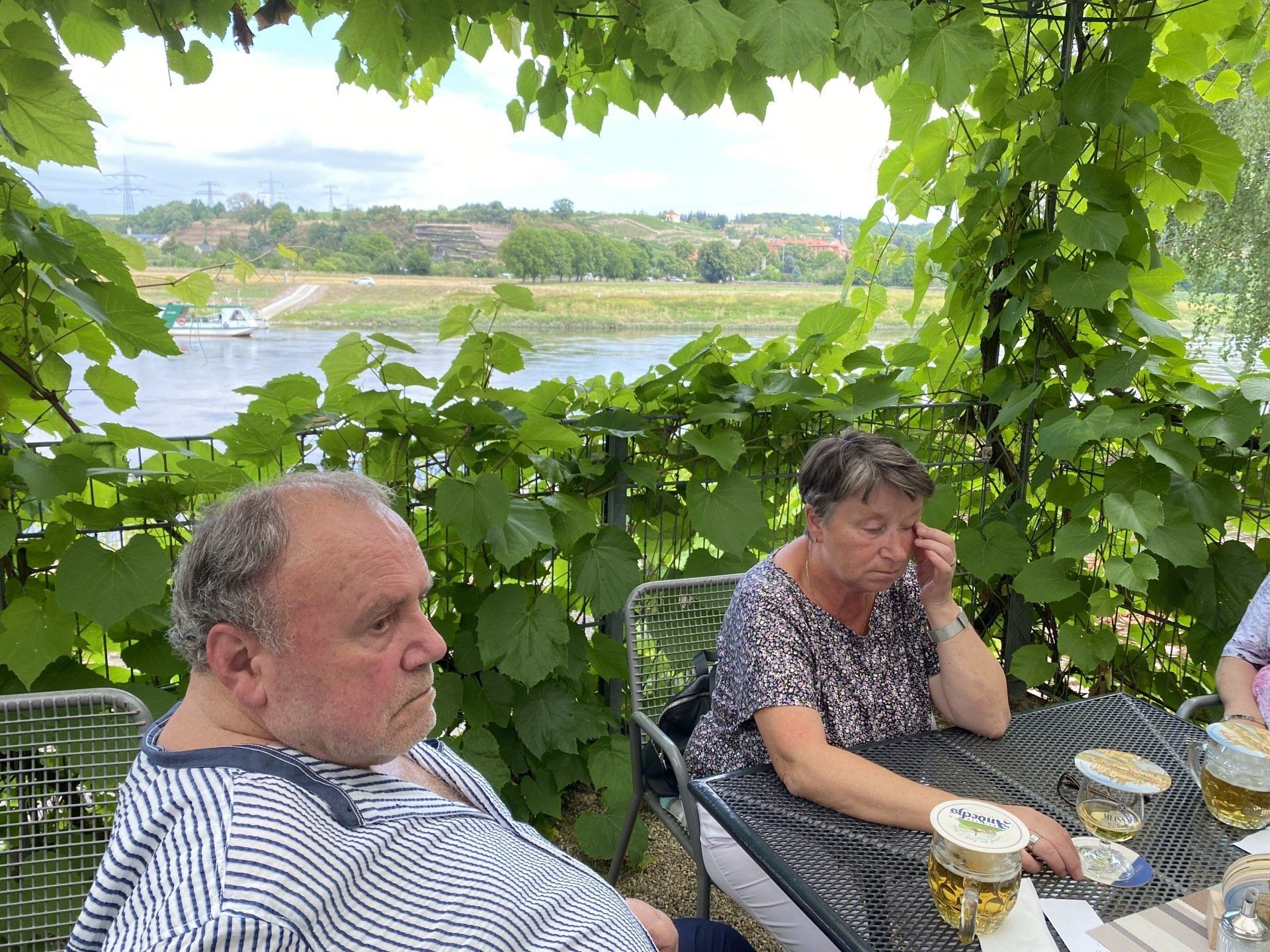 Zwei Personen sitzen an einem Tisch unter einer Weinrebe, im Hintergrund ein Fluss und eine grüne Landschaft.