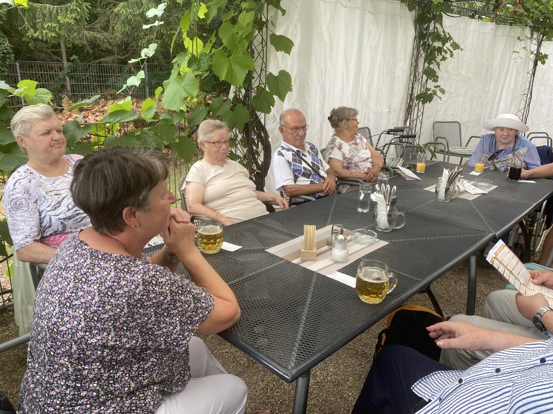 Menschen sitzen an einem Tisch im Freien, einige trinken Bier. Weinblätter hängen über ihnen.