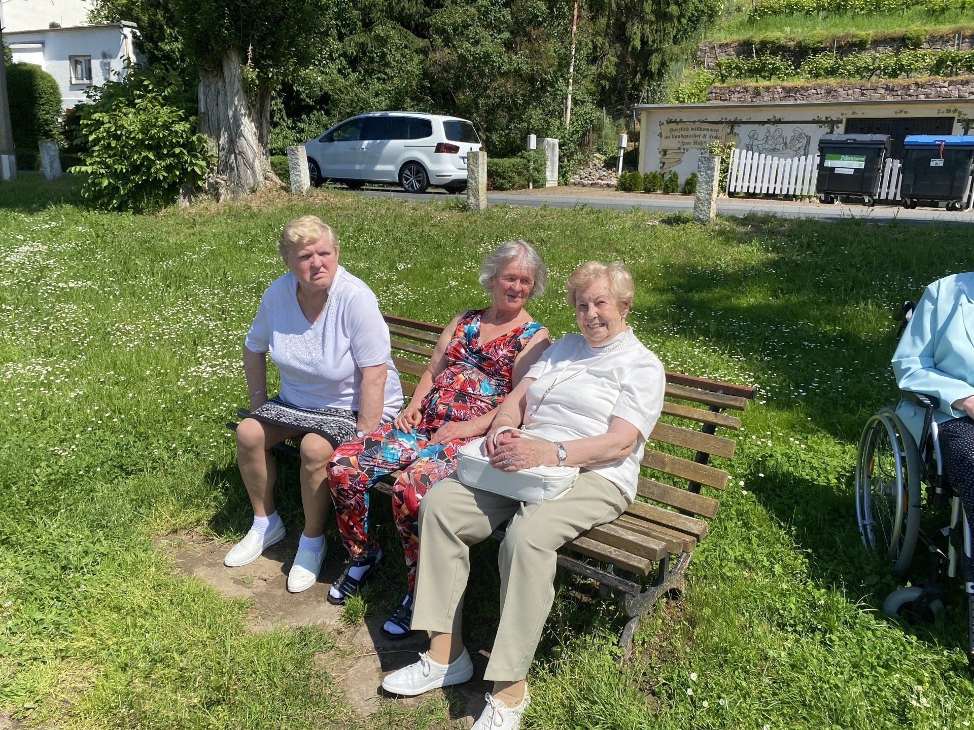 Drei Frauen sitzen auf einer Bank auf einer Rasenfläche, eine weitere sitzt in der Nähe im Rollstuhl. Sonniger Tag.