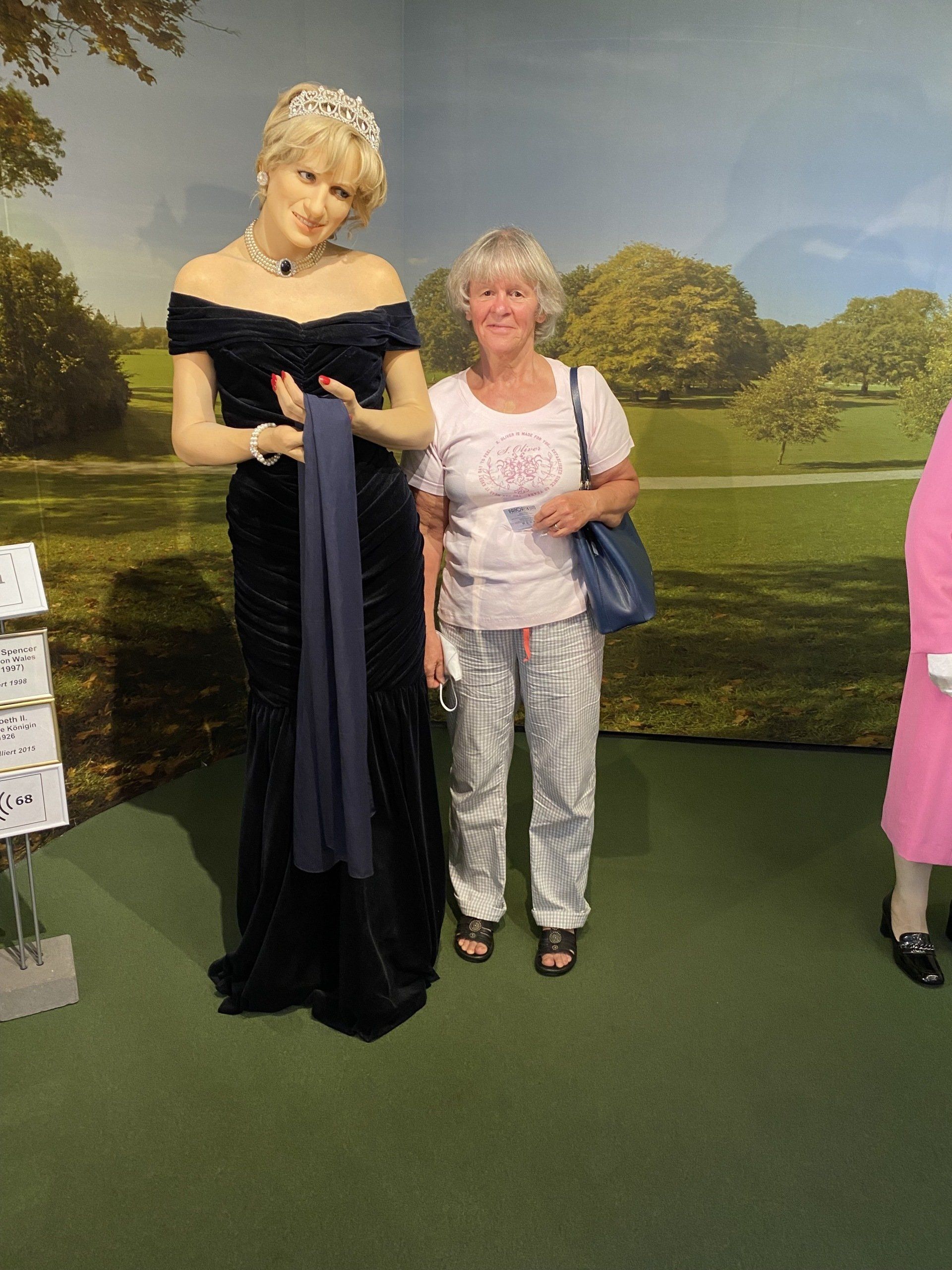 Frau steht neben einer Wachsfigur einer Frau in einem schwarzen Kleid, grüner Teppich, Bäume im Hintergrund.