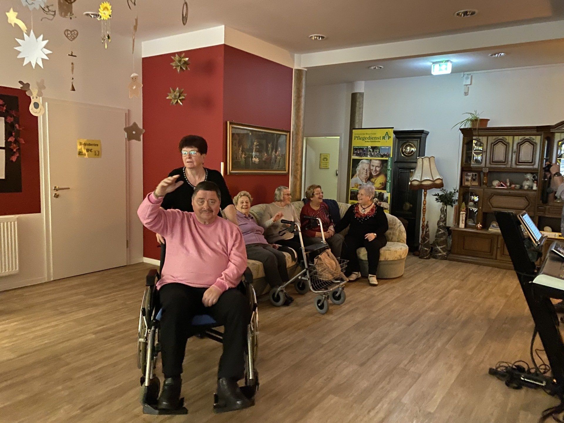 Menschen in einem Wohnzimmer, einige im Rollstuhl, einer winkt. Festliche Dekoration, rote Wand, Holzboden.