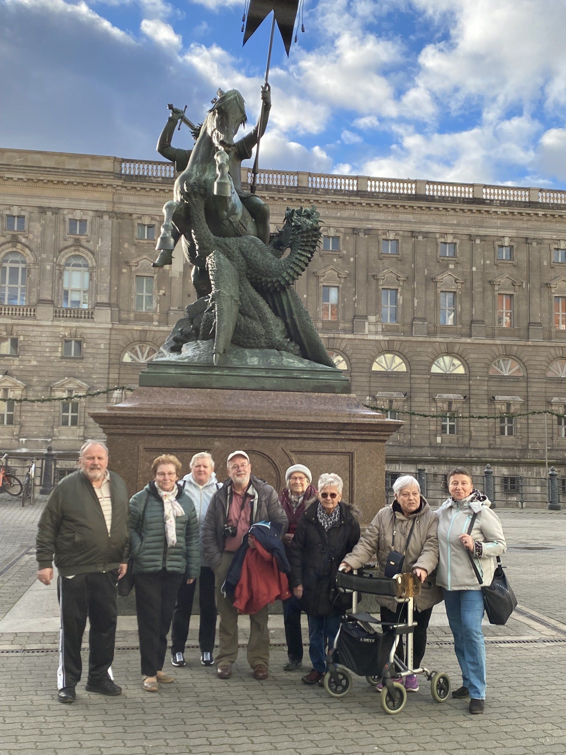 Eine Gruppe von Menschen posiert vor der Statue eines Drachentöters und einem großen Gebäude. Bewölkter Himmel.
