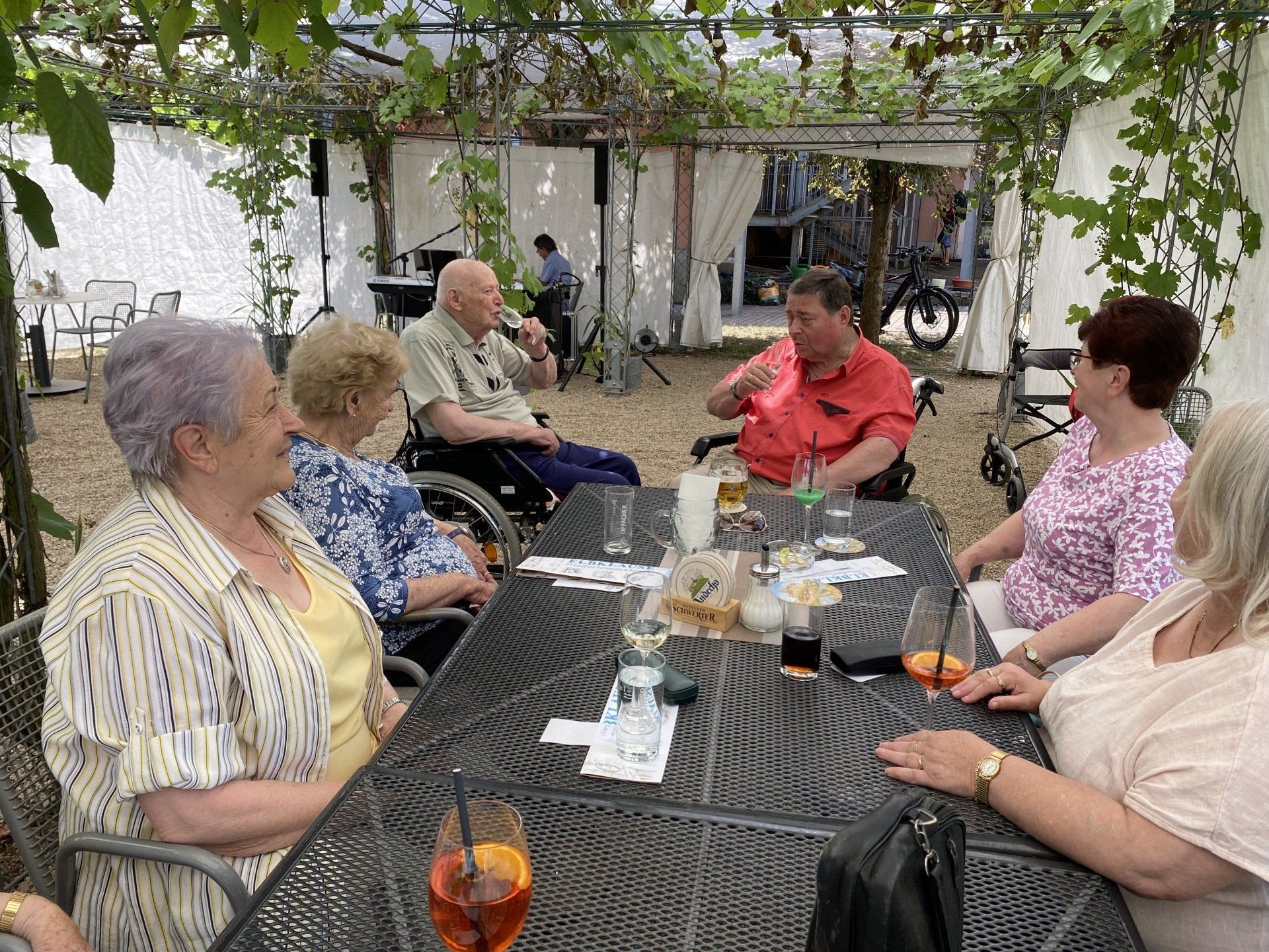 Eine Gruppe von Menschen im Rollstuhl sitzt an einem Tisch im Freien, unterhält sich und trinkt.