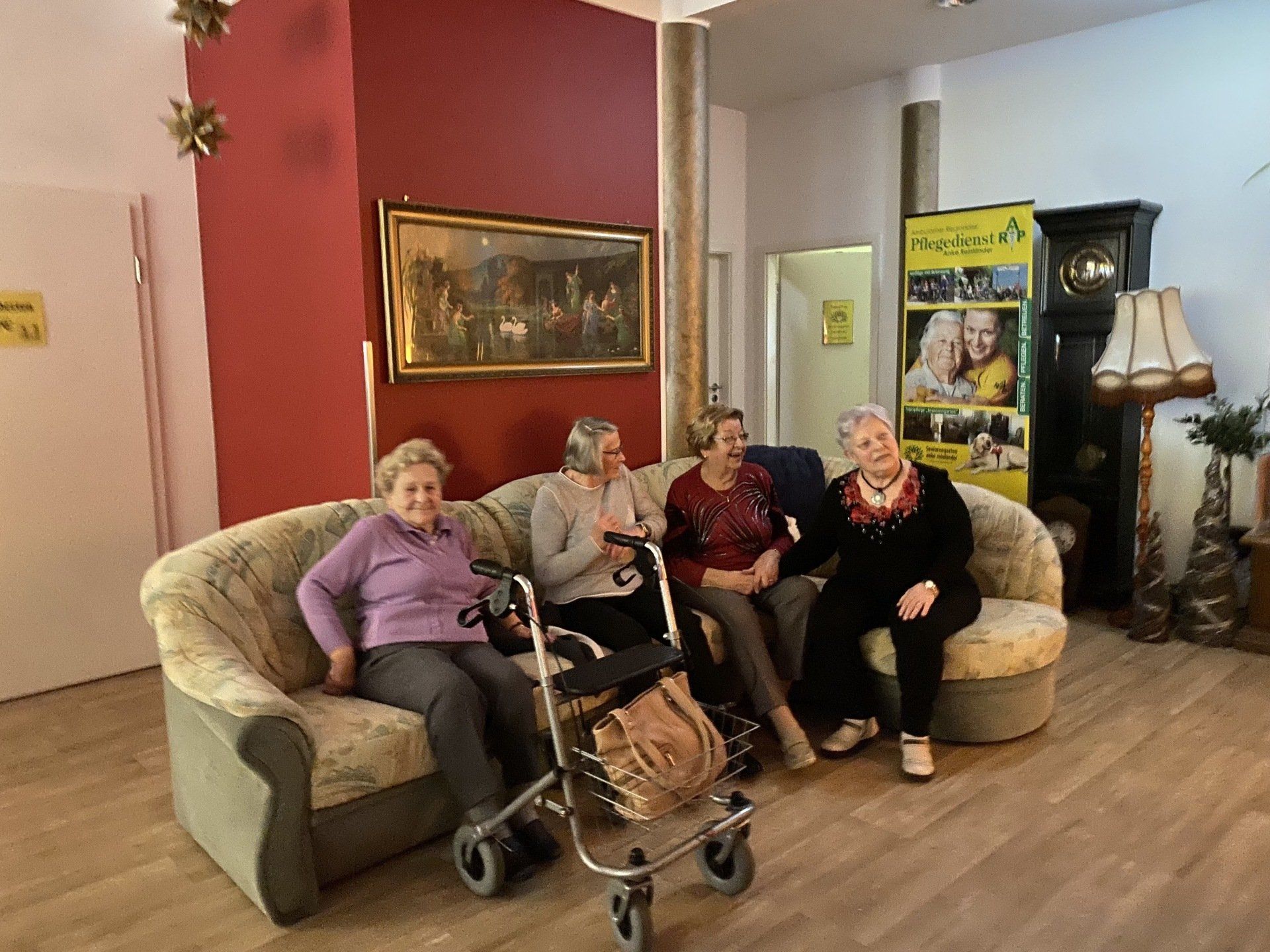 Vier Personen sitzen auf einem Sofa in einem hell erleuchteten Raum. Eine Person trägt einen Rollator.