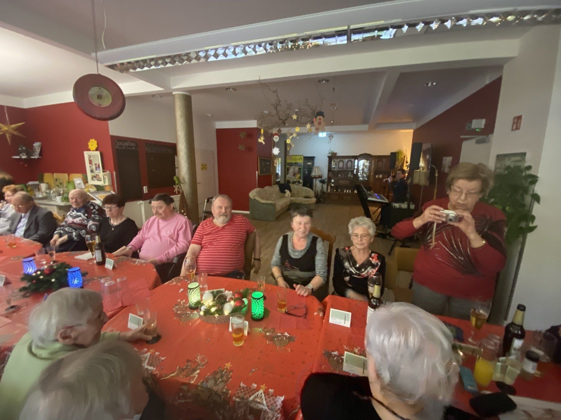 Menschen sitzen um einen langen roten Tisch. Eine Person macht ein Foto. Festliche Atmosphäre mit Dekorationen und Getränken.
