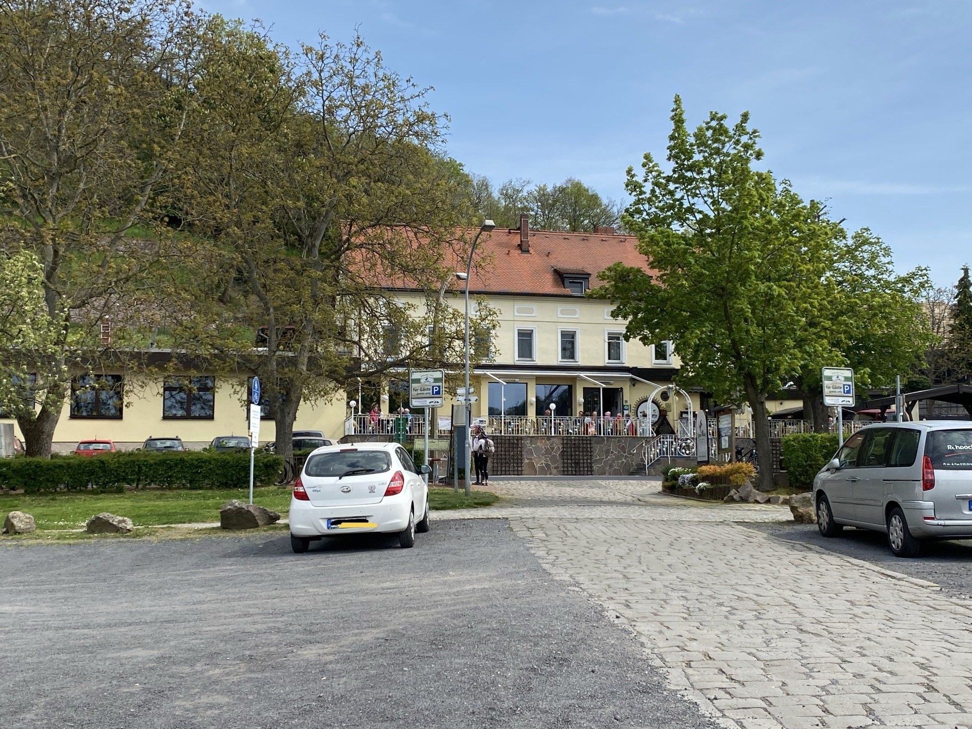Außenbereich des Restaurants mit Sitzgelegenheiten im Freien, Parkplatz, Bäumen und Autos.