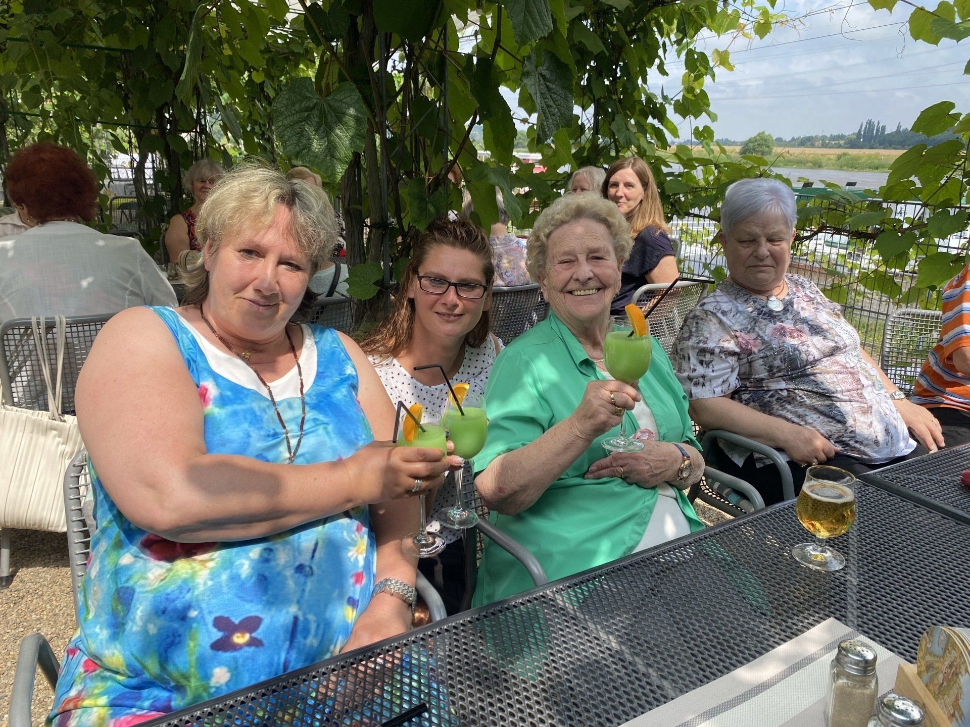 Vier Frauen an einem Tisch im Freien stoßen mit grünen Getränken an. Im Schatten der Pflanzen, sonniger Tag.