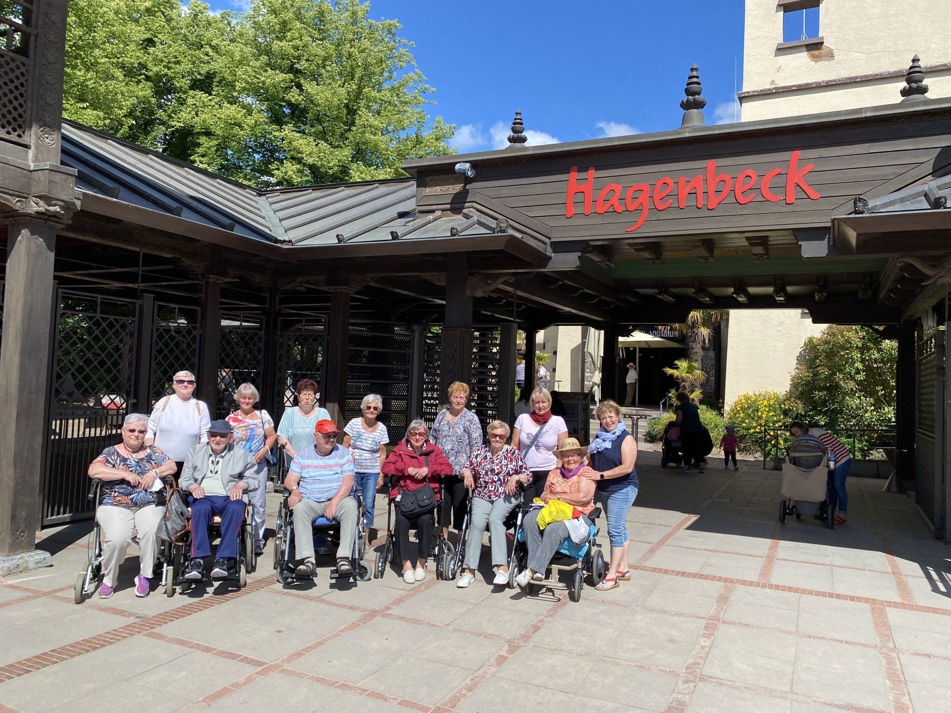 Gruppe von Menschen im Rollstuhl vor einem Gebäude mit Torbogen. Oben Schild „Hirscheneck“.