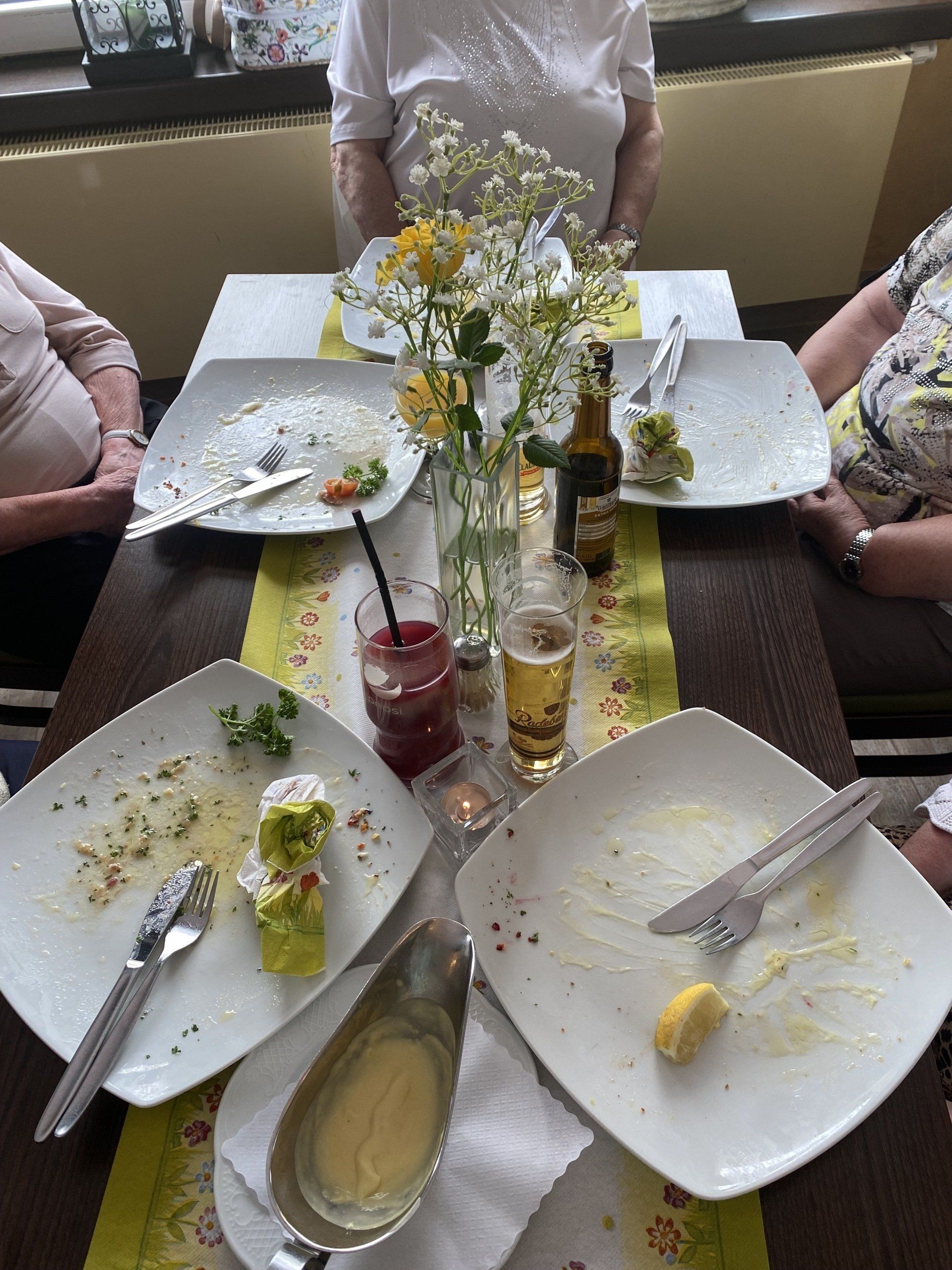 Leere Teller auf einem Restauranttisch mit Essensresten, einer Sauciere, Getränken und sitzenden Personen.