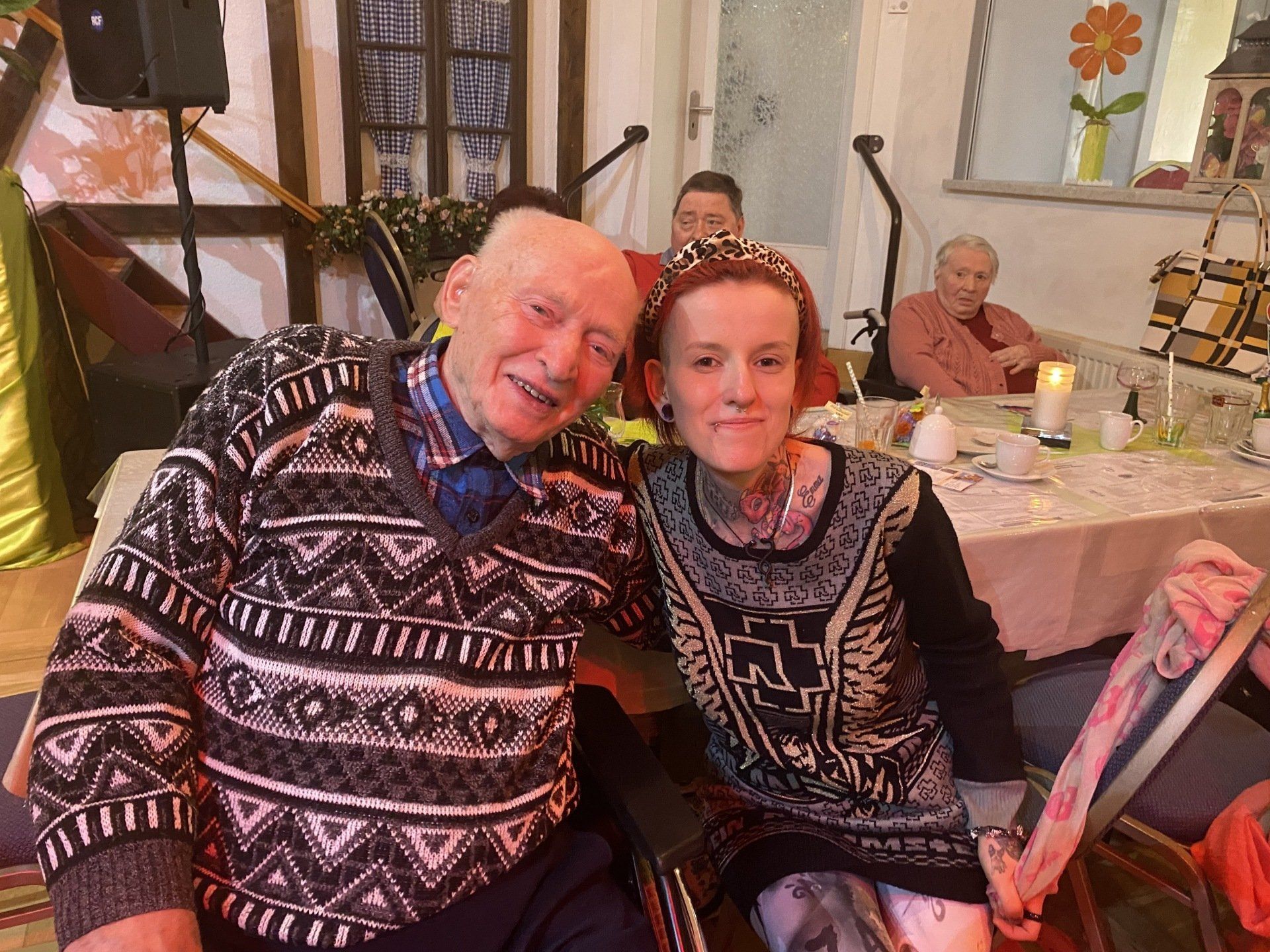 Lächelnde Menschen in einem Raum, einer in einem gemusterten Pullover, sitzen neben einem dekorierten Tisch.
