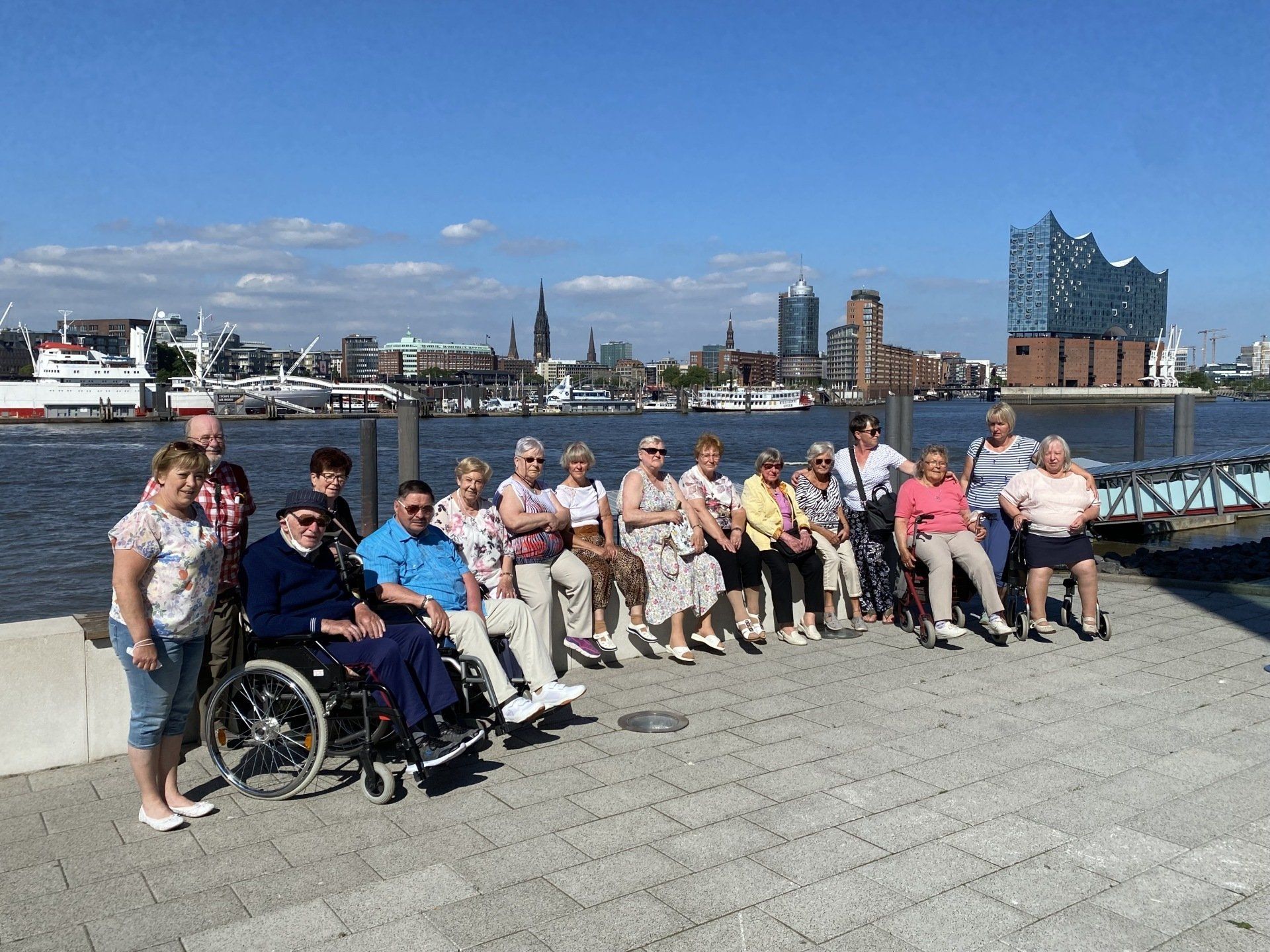 Eine Gruppe von Menschen, einige davon im Rollstuhl, posiert im Freien am Wasser vor der Skyline der Stadt im Hintergrund.