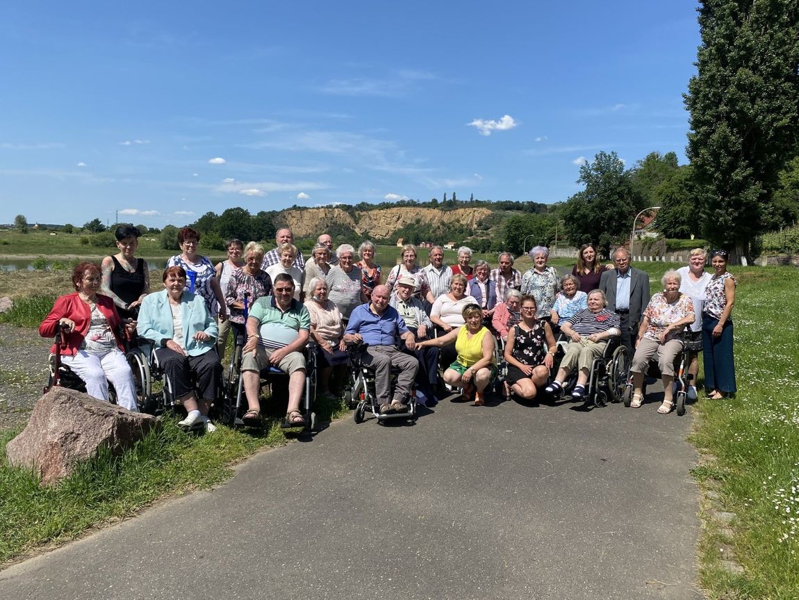 Gruppe von Menschen posiert im Freien; einige im Rollstuhl, sonniger Tag mit blauem Himmel.