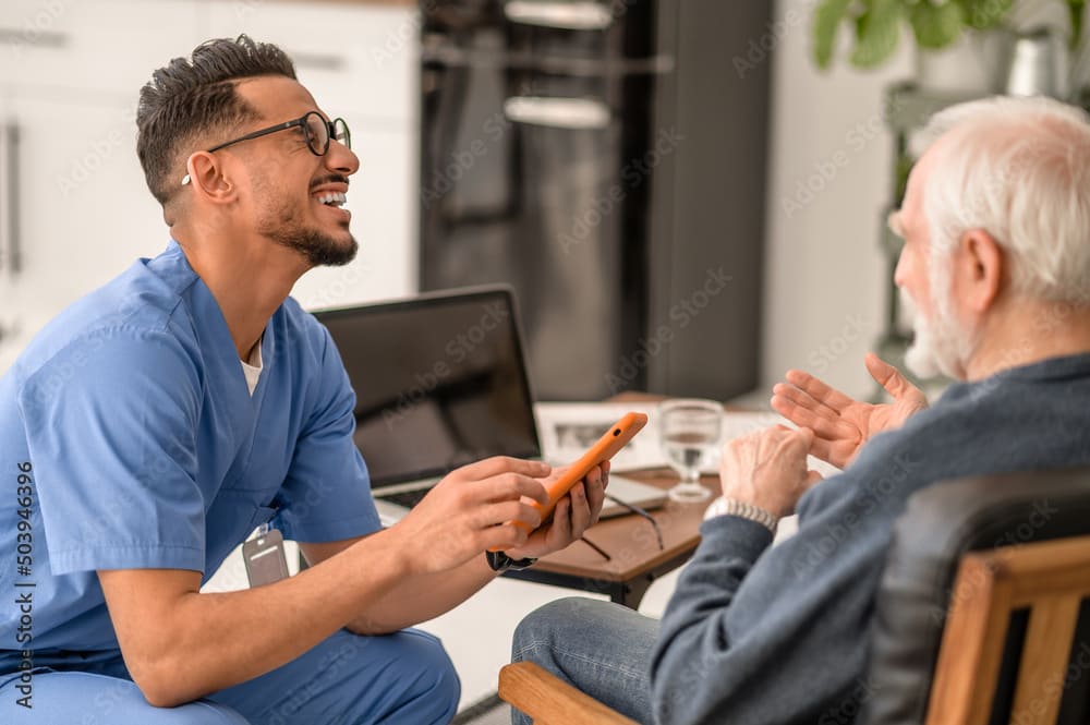Eine Pflegekraft im blauen Kittel lächelt, während sie einem älteren Mann ein Tablet zeigt.