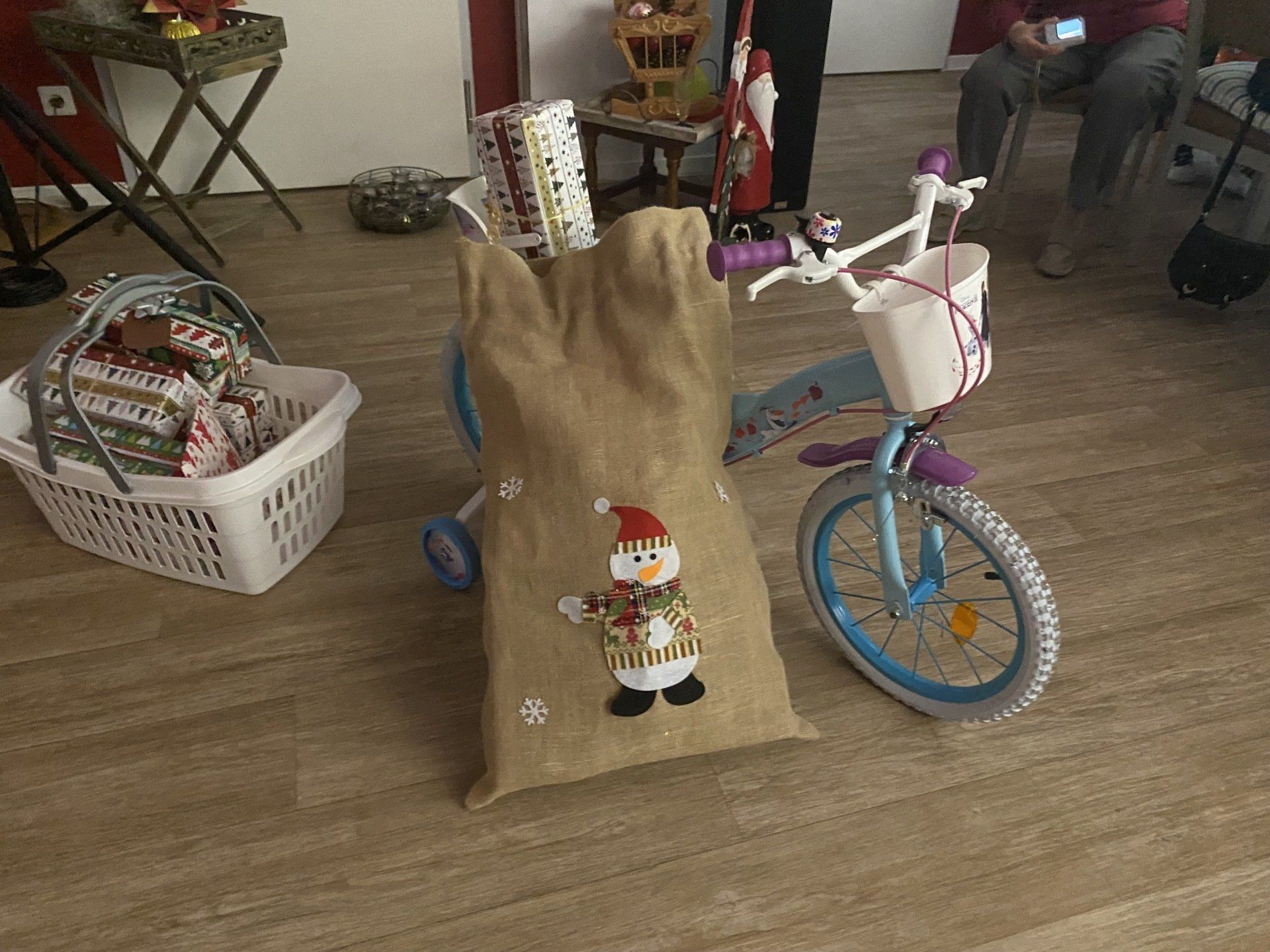 Weihnachtssack mit Schneemann darauf neben einem blauen Kinderfahrrad und Geschenken in einem Korb.