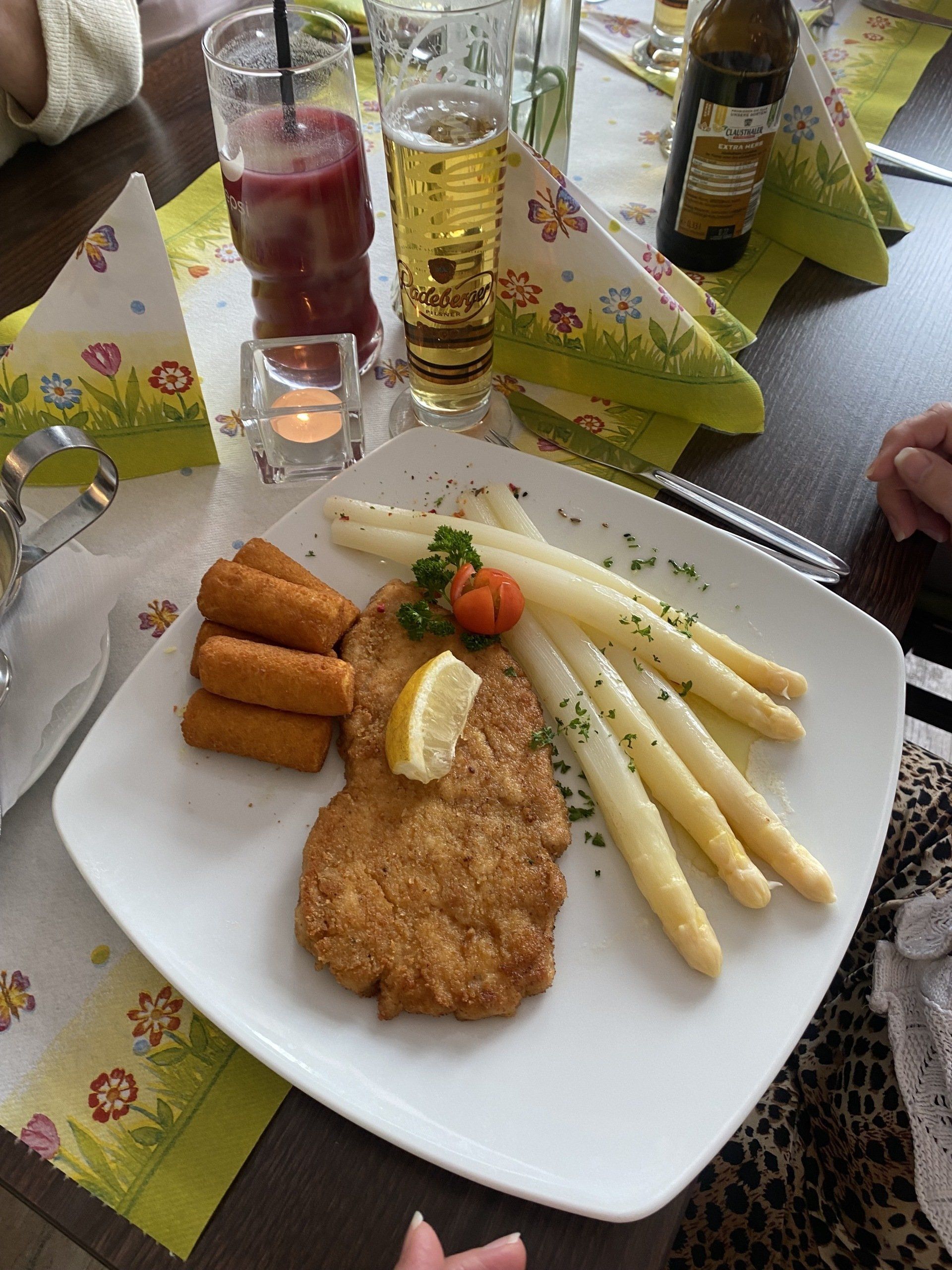 Teller mit Essen: paniertes Fleisch, Spargel, Kroketten, Tomaten, Zitronenscheibe. Getränke und florale Tischdekoration.