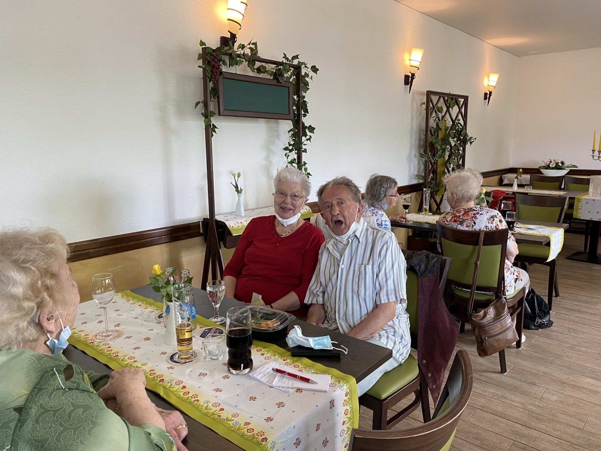 Menschen an Tischen in einem Restaurant. Zwei lächeln, andere im Hintergrund. Mit Pflanzen dekoriert.