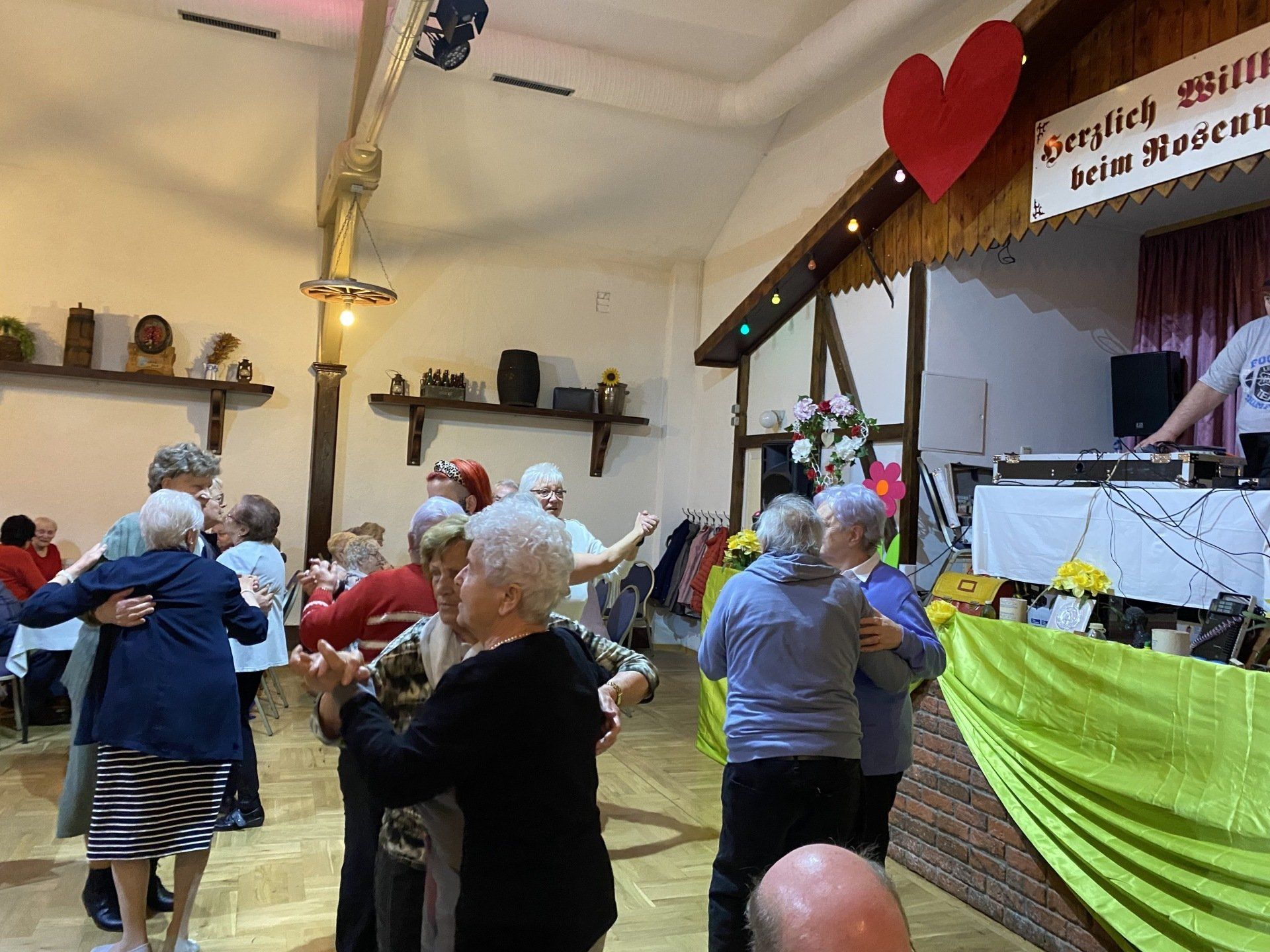 Senioren tanzen in einem Saal; DJ, Bühne mit Herzdeko, Holzbalken, diverse Dekorationen.