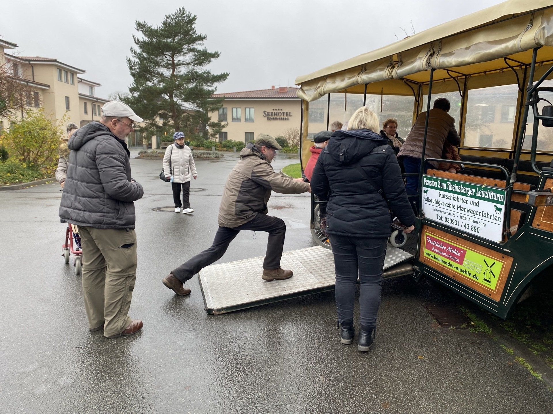Menschen steigen in einen Trolley mit Rampe ein. Ein Mann hilft jemandem, andere warten auf dem Trolley oder daneben.