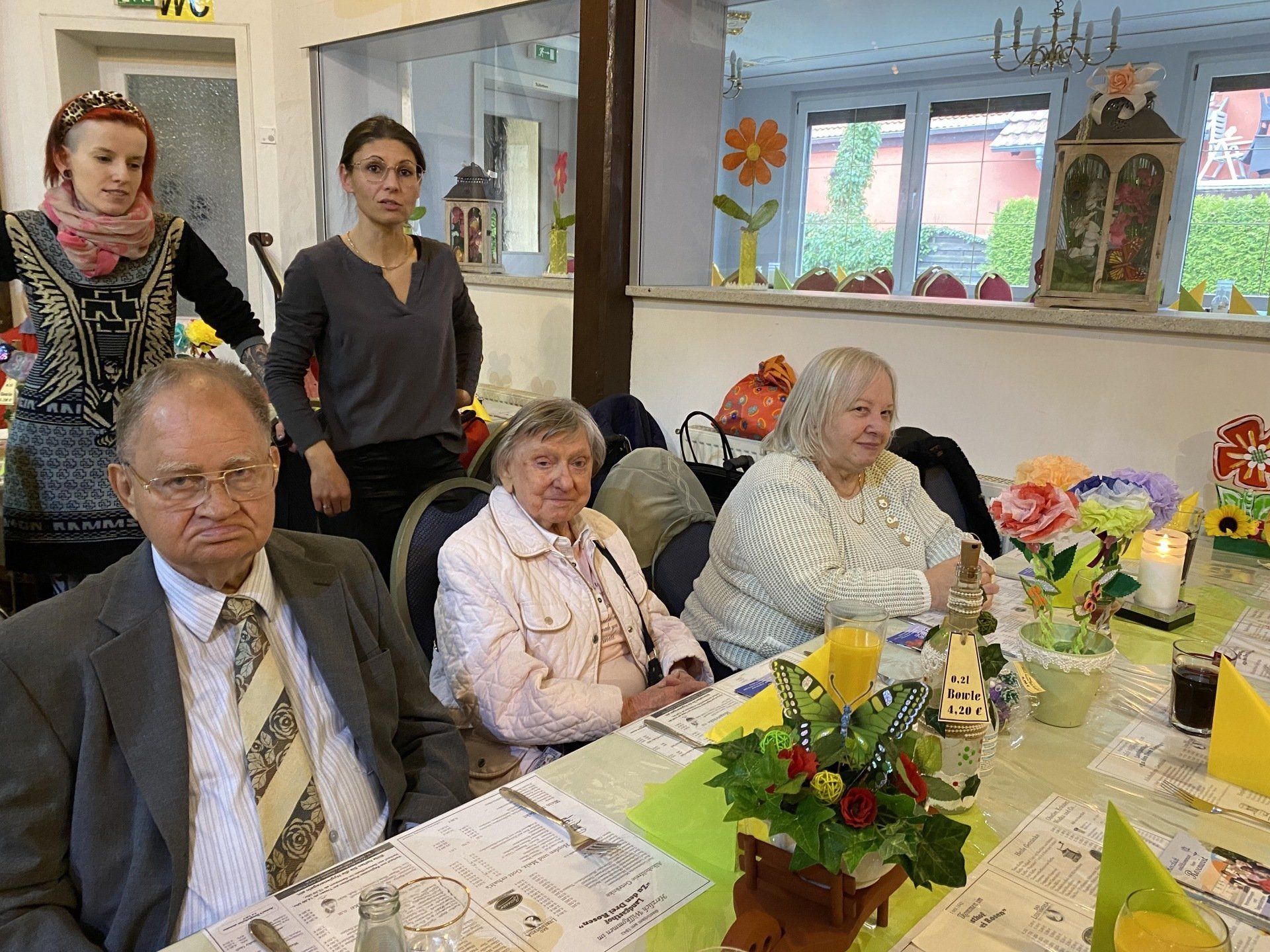 Menschen an einem Tisch in einem dekorierten Raum. Eine Frau vorn, zwei stehen hinter den sitzenden Menschen.