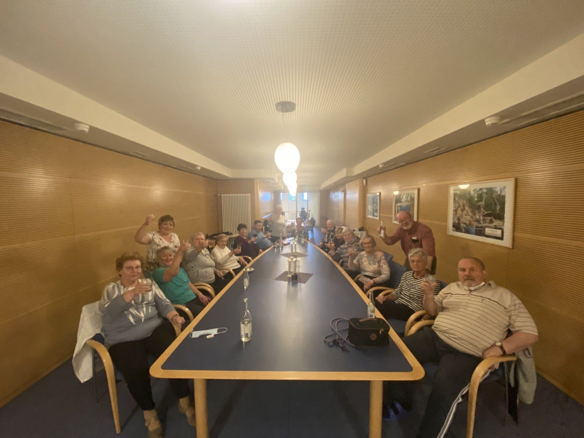 Menschen sitzen um einen langen Tisch in einem holzgetäfelten Raum. Einige winken.