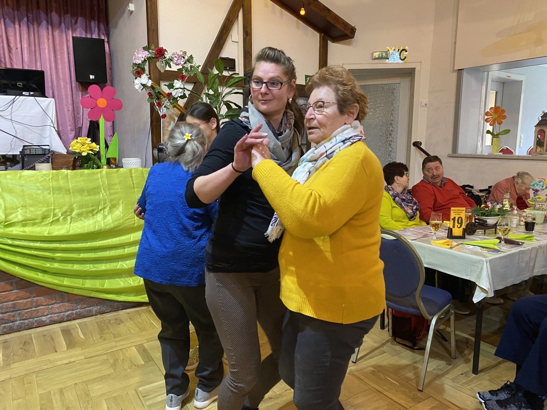 Zwei Frauen tanzen in einem Saal. Eine trägt einen gelben Pullover, die andere ein schwarzes Hemd. 