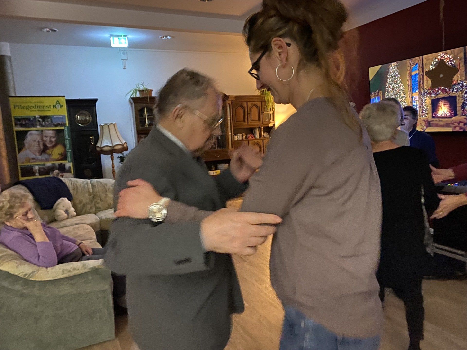 Frau tanzt mit einem Mann im Innenbereich. Andere schauen zu. Warm beleuchteter Raum mit Möbeln und Dekorationen.