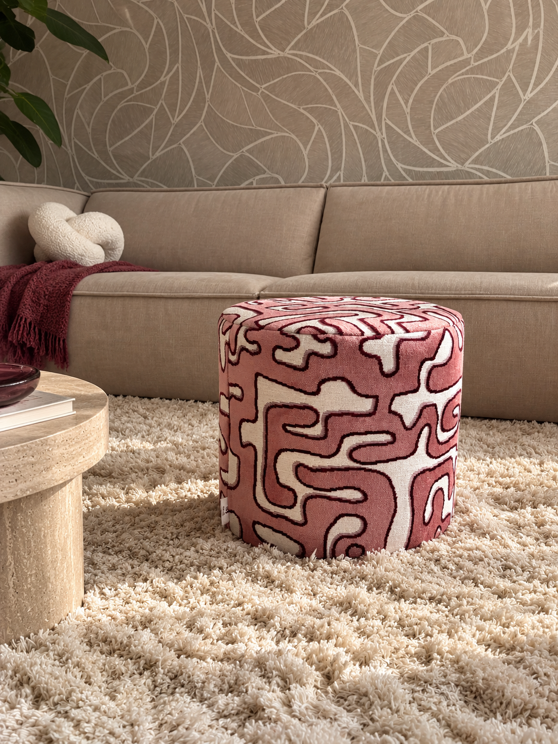 Roze poef met patroon op een hoogpolig vloerkleed in een woonkamer met een beige bank en behang als achtergrond