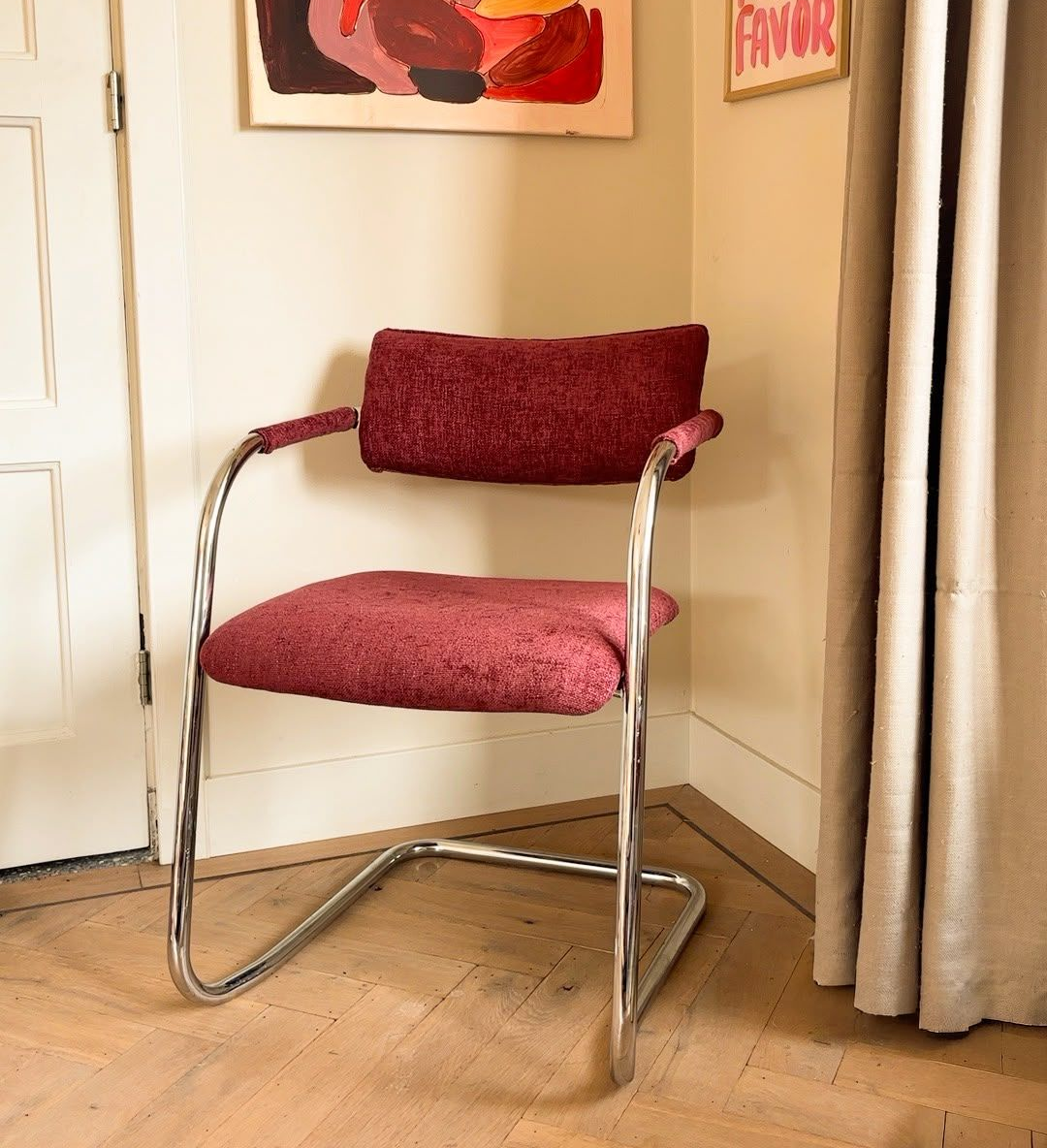 Bordeauxrode fauteuil met bekleding, een chromen frame, in een hoek naast gordijnen en wanddecoratie