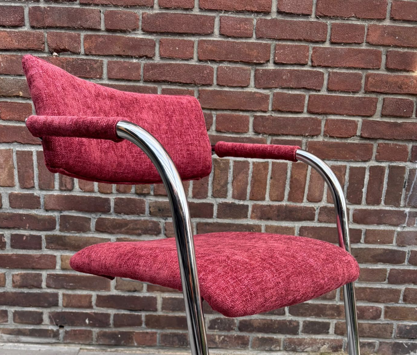 Rood gestoffeerde stoel met chromen armleuningen tegen een bakstenen muur
