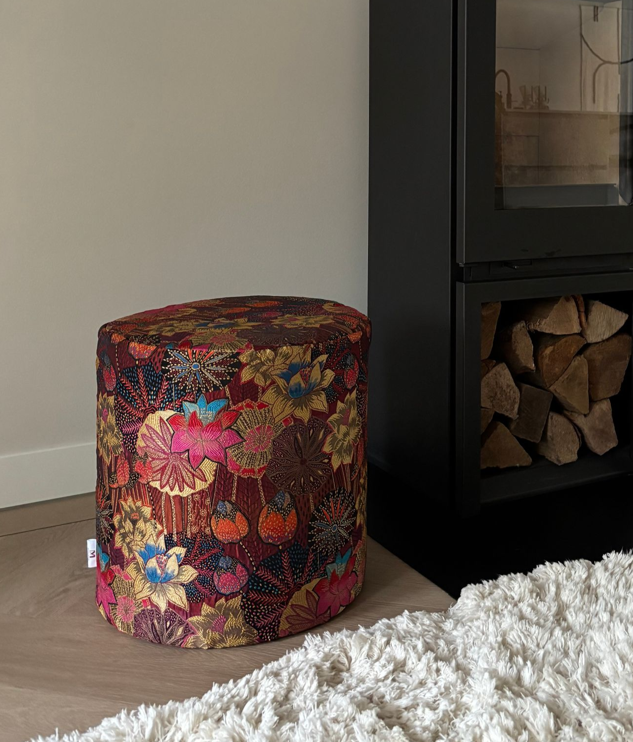 Kleurrijke poef met bloemenprint naast een zwarte open haard in een woonkamer.