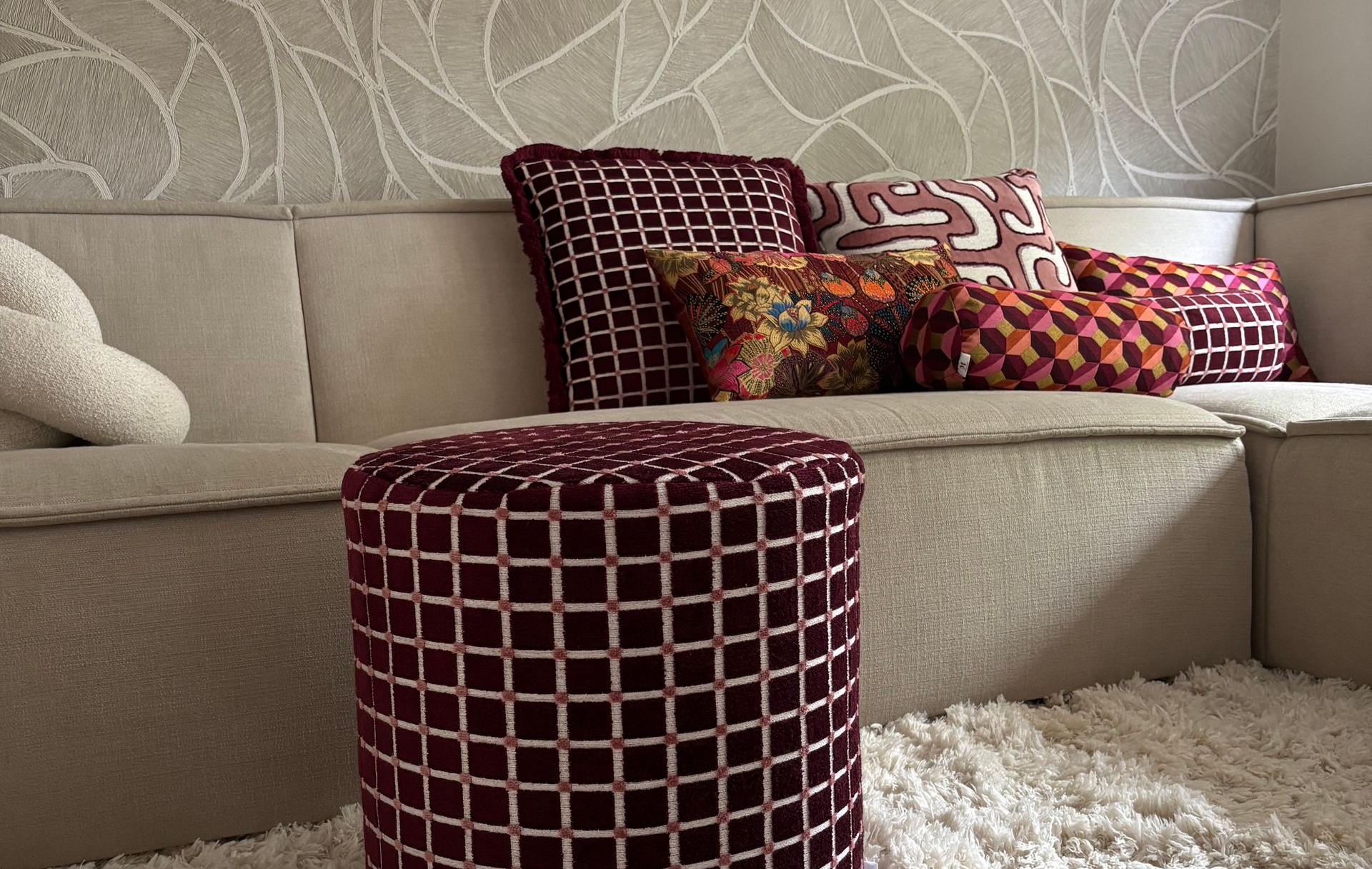 Beige hoekbank met sierkussens, een bordeauxrode poef en een wit hoogpolig vloerkleed in een moderne woonkamer.