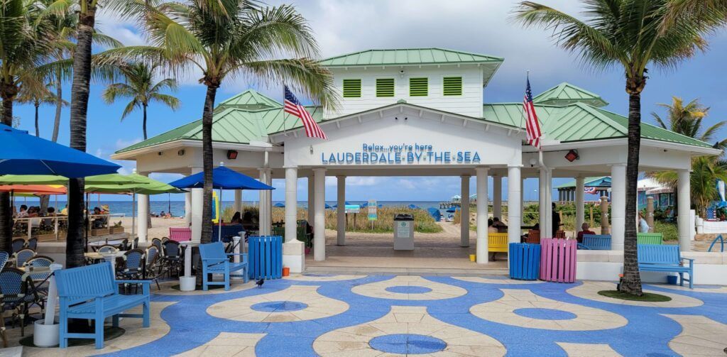 Beachside entrance with palm trees, blue patterned pavement, and white-green pavilion under a clear sky