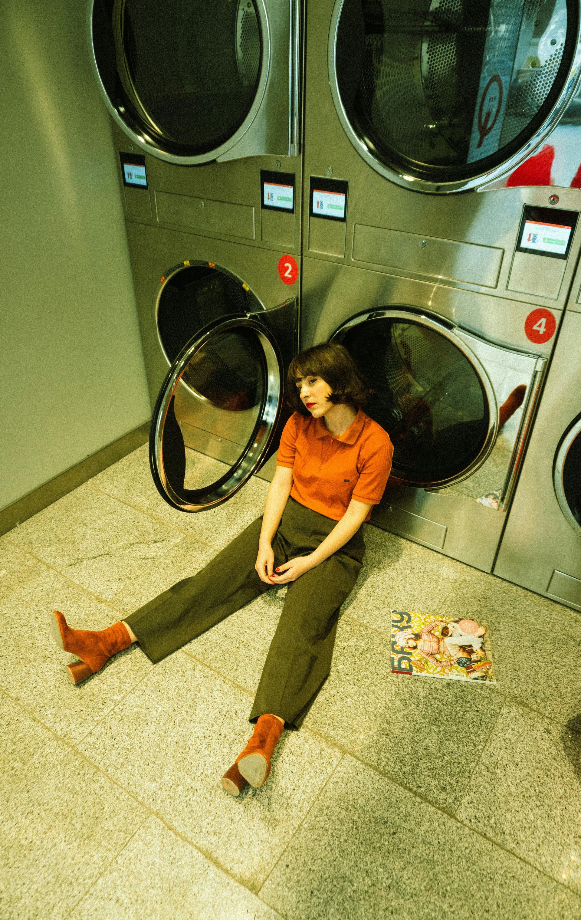 Person in orange shirt sitting on a laundry room floor beside open dryer doors and a pile of clothes.