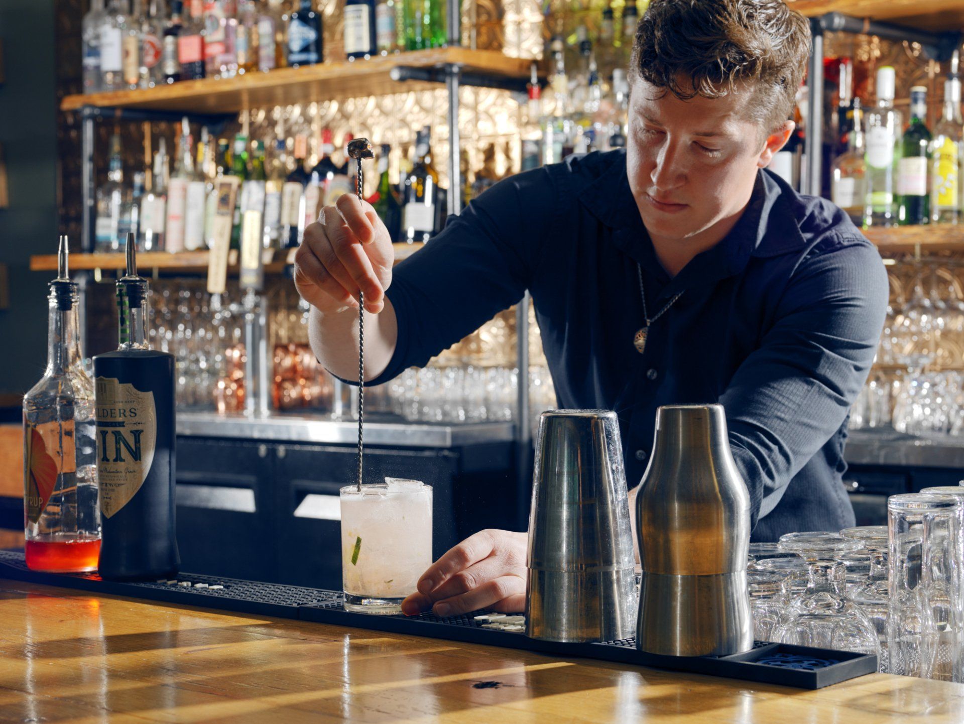 Bartender stirring a cocktail behind a bar, with bottles and shakers in the background.