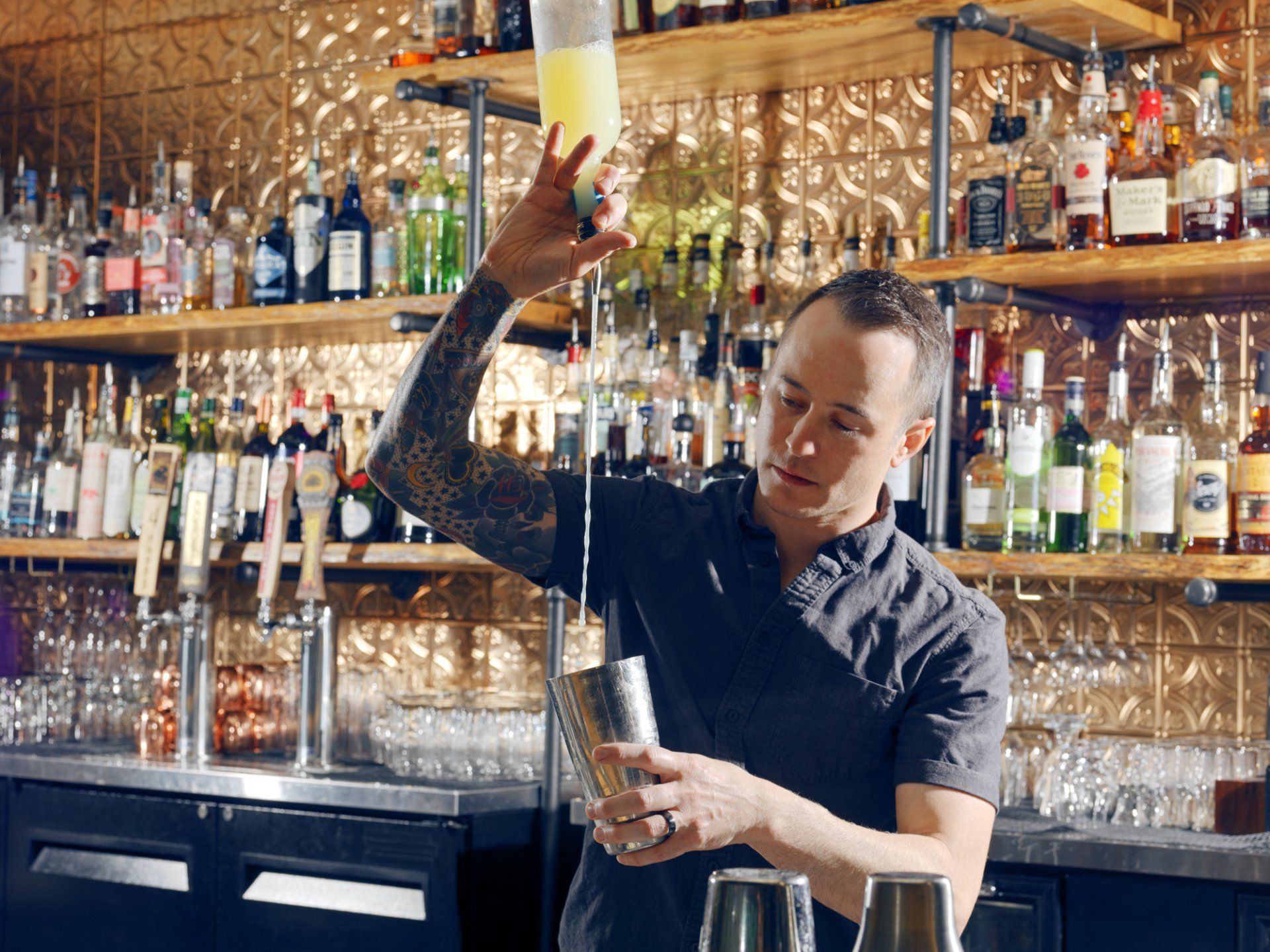 Bartender pouring a yellow cocktail into a shaker behind a stocked bar with hanging glasses