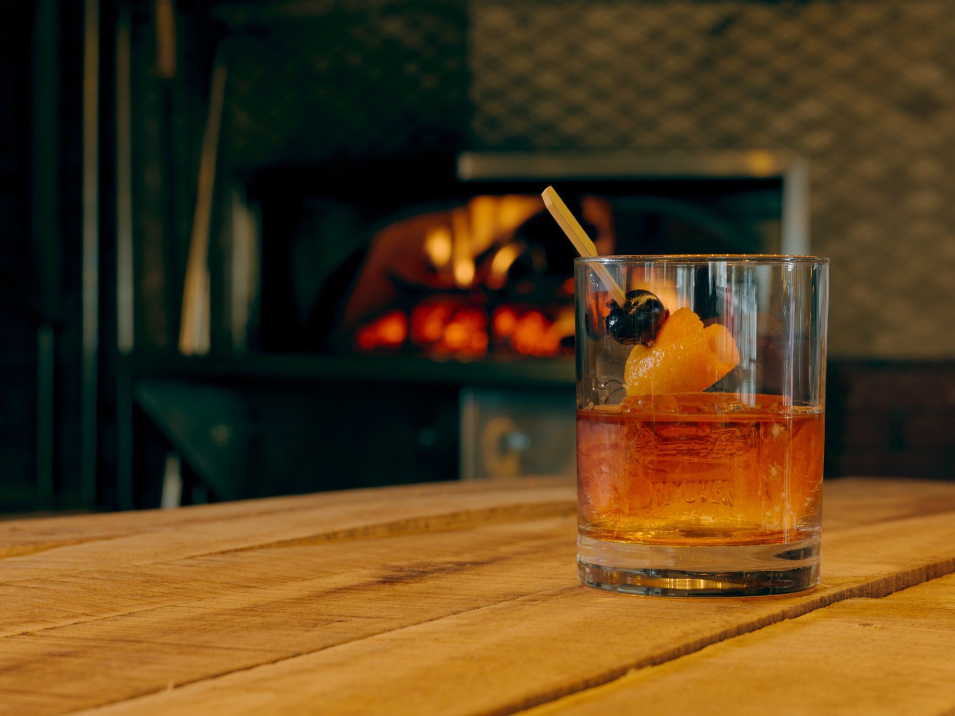 Whiskey cocktail with orange peel and cherry on a wooden table by a fireplace