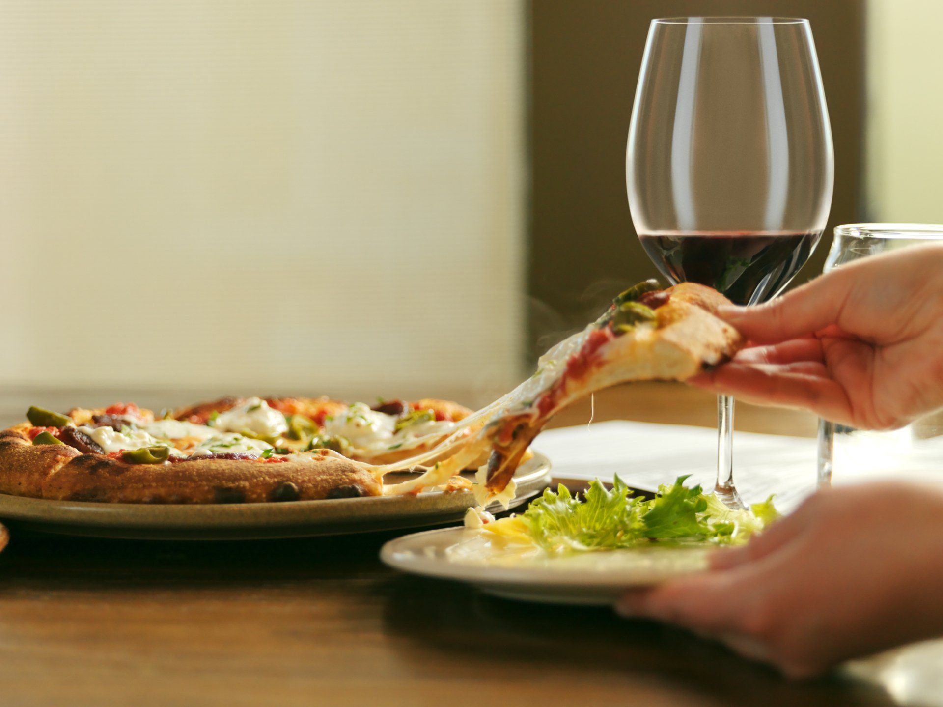 Hand taking a slice of pizza beside a wine glass and salad on a dining table