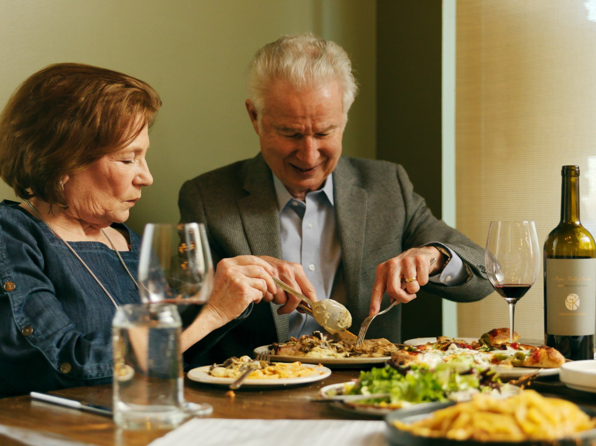 Older couple dining at a table, serving food and smiling in a warm restaurant setting.