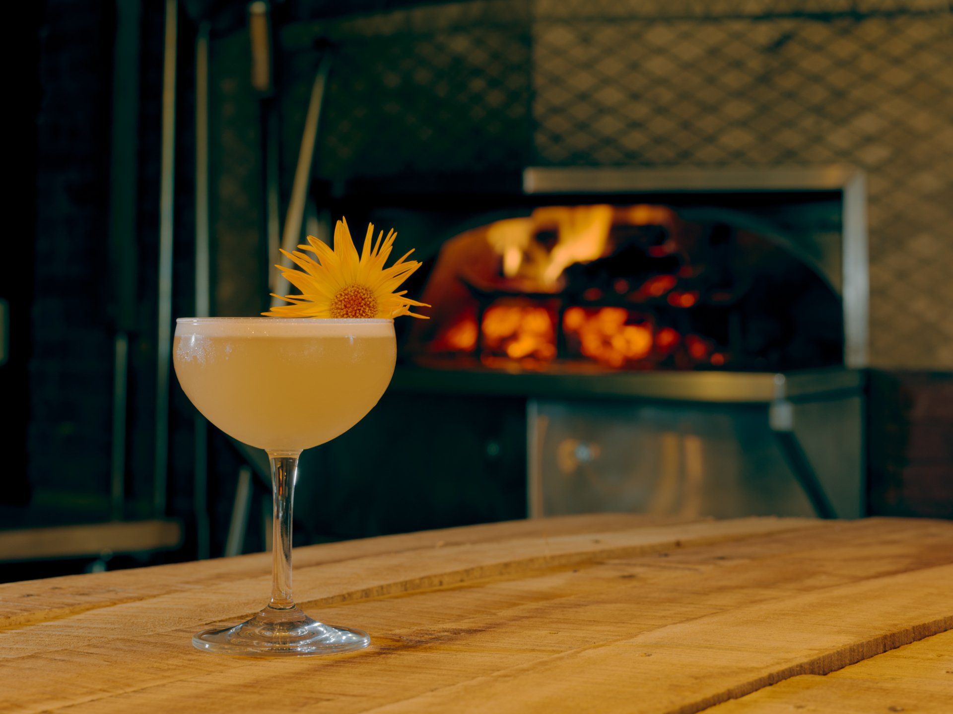 Cocktail in a coupe glass on a wooden table, with a lit fireplace in the background