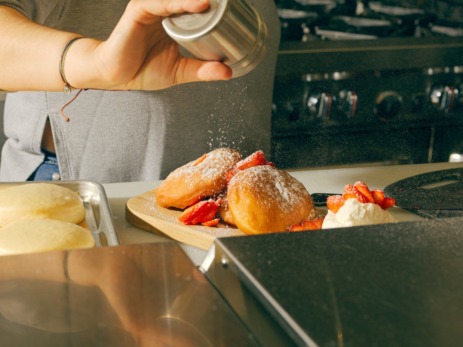 Hand dusting powdered sugar over donuts on a kitchen counter beside a stove