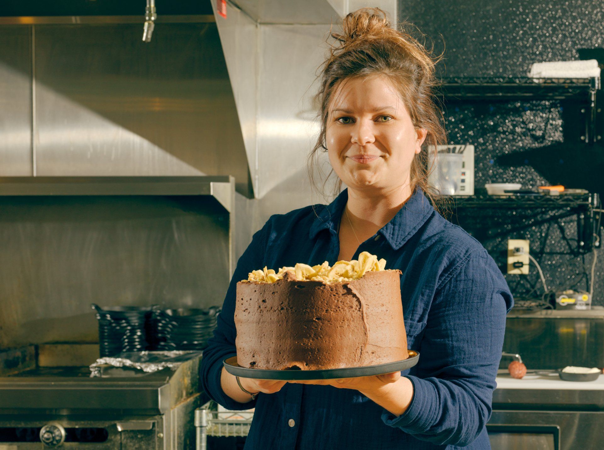Person in a blue shirt holding a chocolate cake in a kitchen
