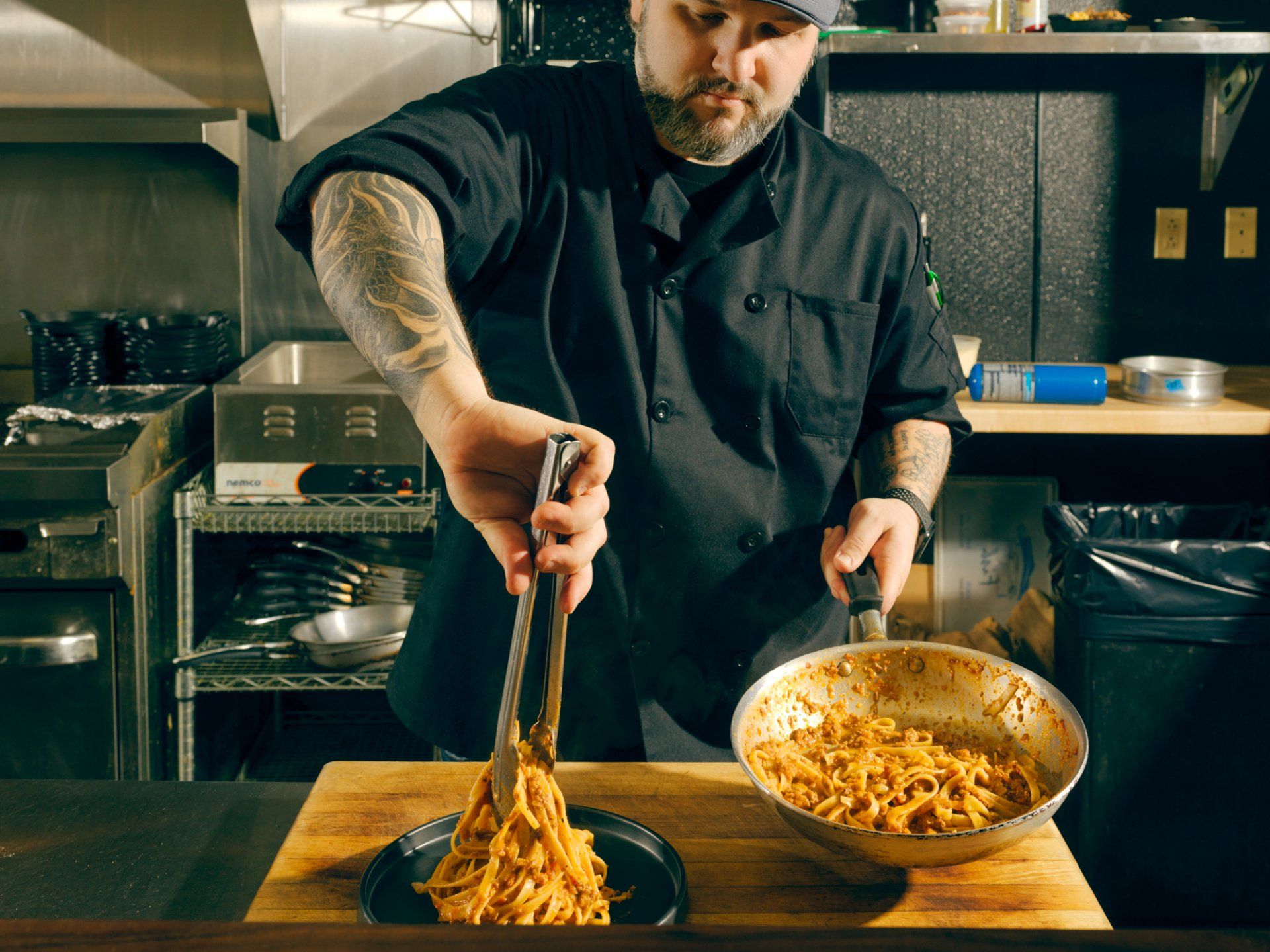 Chef plating pasta in a restaurant kitchen with a large bowl of sauce beside him