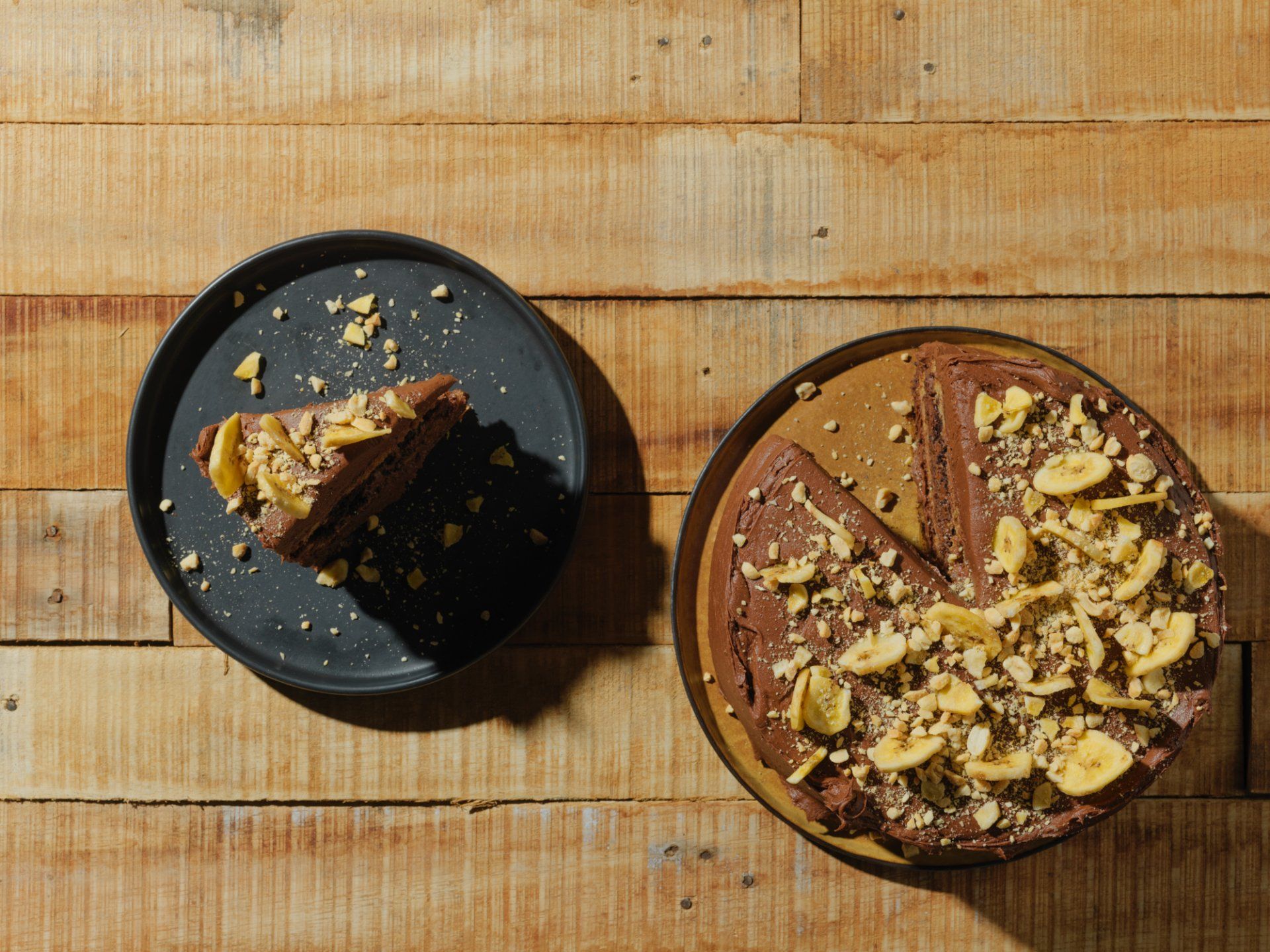 Brown cake on a brown plate on a wooden table, topped with chopped nuts and a slice removed on a black plate