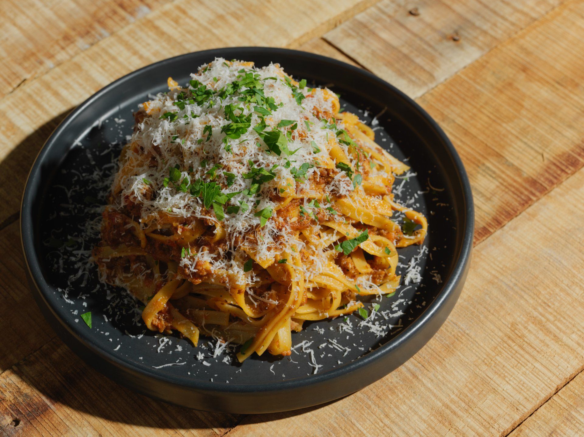 Black bowl of pasta topped with grated cheese and herbs on a wooden table