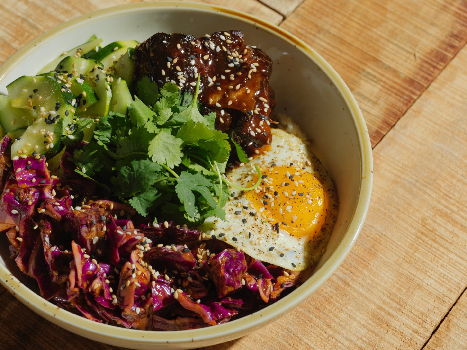 Bowl of rice with fried egg, avocado, cabbage, cilantro, and sauce on a wooden table