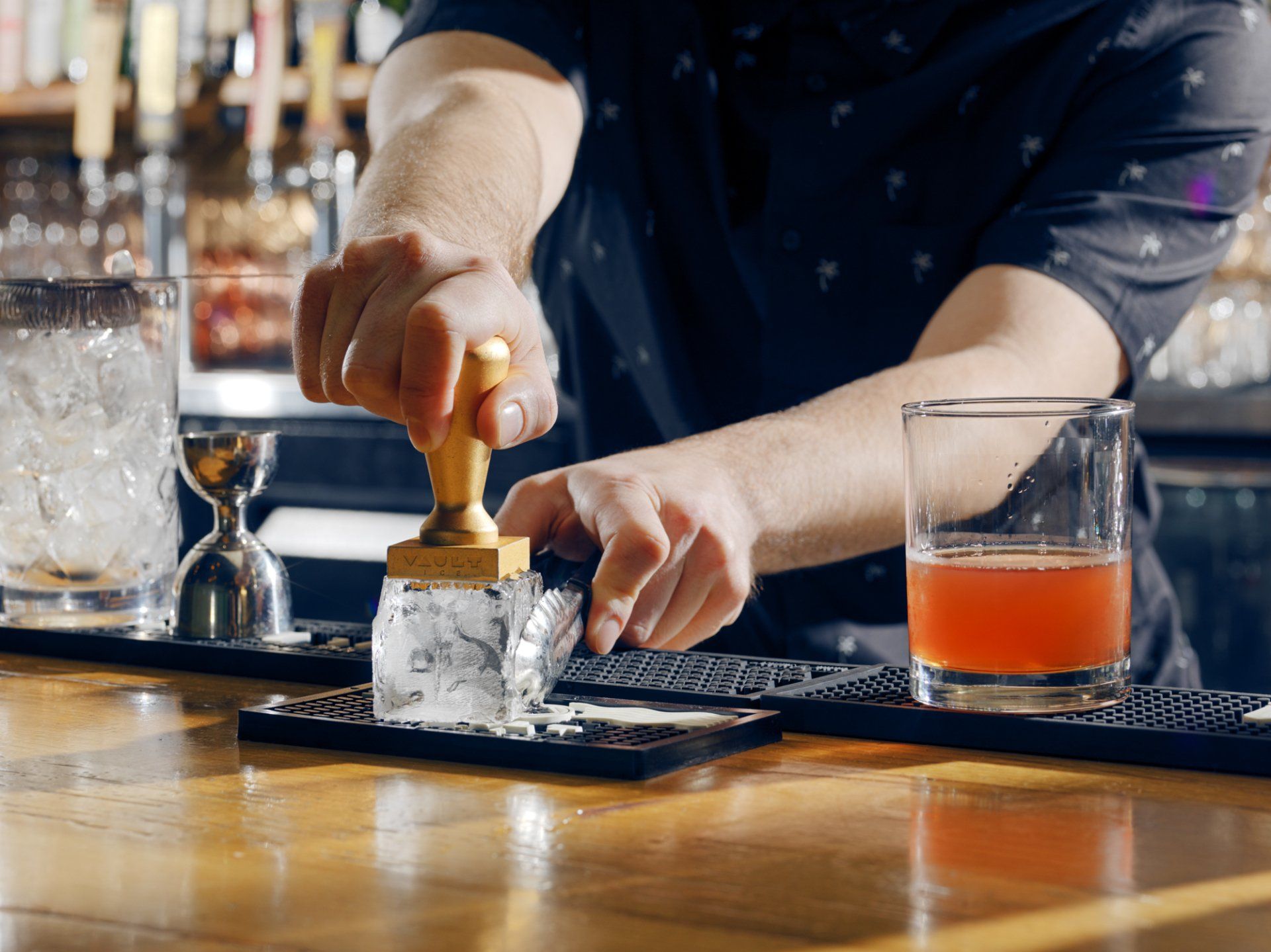 Bartender pressing a drink into ice with a wooden muddler beside two cocktails on a bar counter