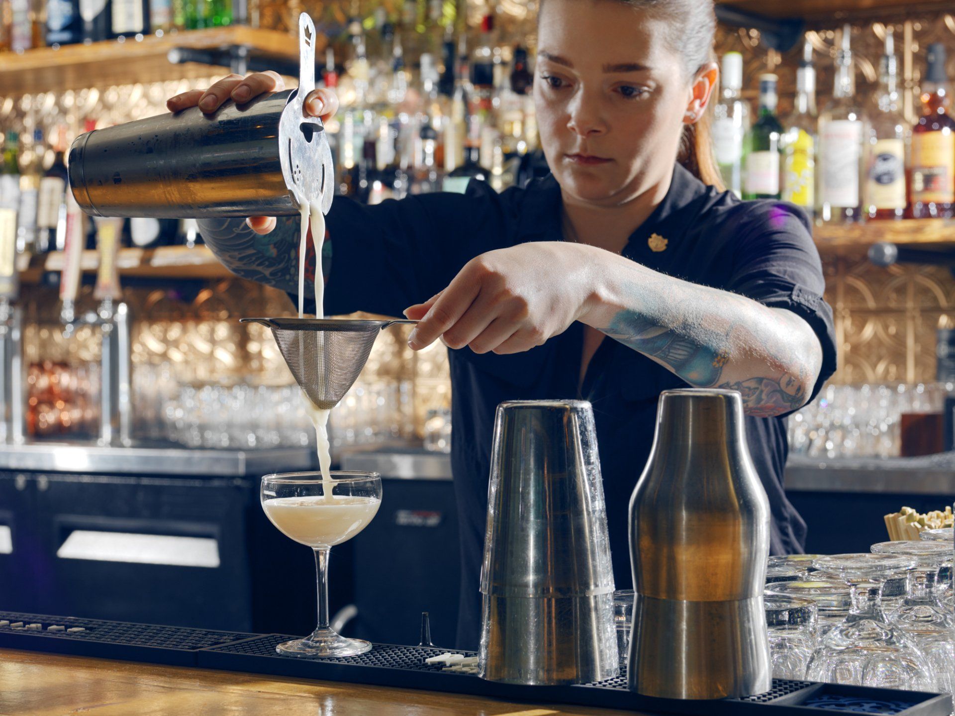 Bartender straining a cocktail into a glass behind a bar, with bottles and shakers in the background