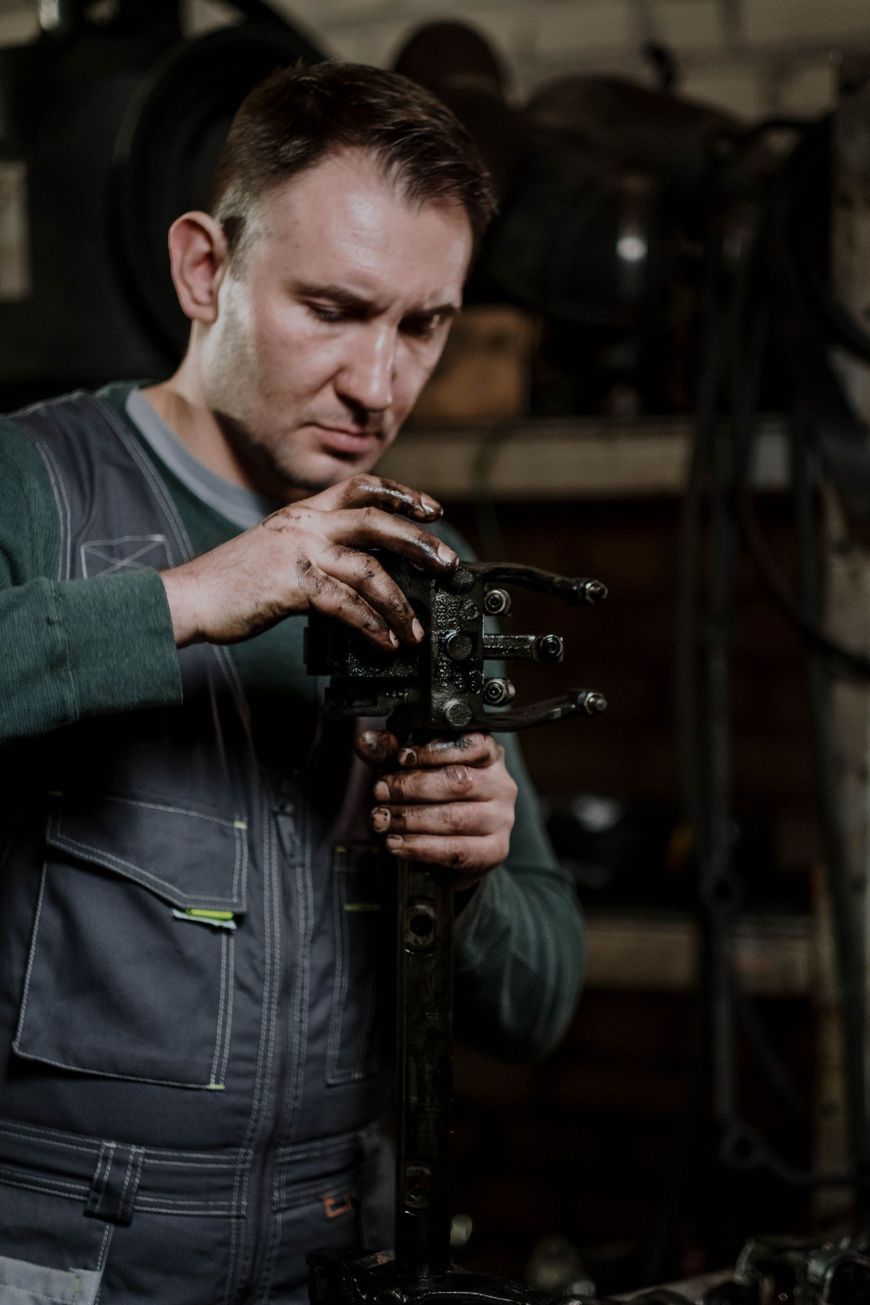 Mechanic inspecting greasy car part in a dim workshop, wearing gray overalls