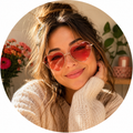 Smiling person in heart-shaped pink sunglasses and a cream sweater, resting their chin in a warm indoor setting